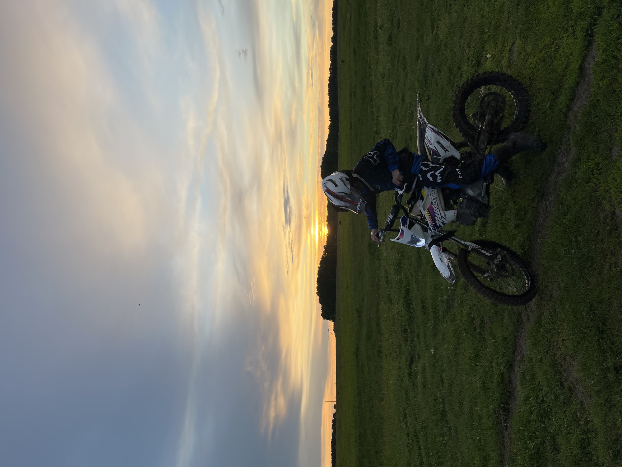 A person wearing a helmet and blue riding gear is seated on a white dirt bike in a grassy field. The dirt bike is angled towards the right side of the image. The background features a sunset with orange and yellow hues blending with the sky. A line of trees is visible along the horizon. The photo appears to be taken in a rural setting.
Based on visual elements, the location in the city of Новочувашский, Russia cannot be determined.
