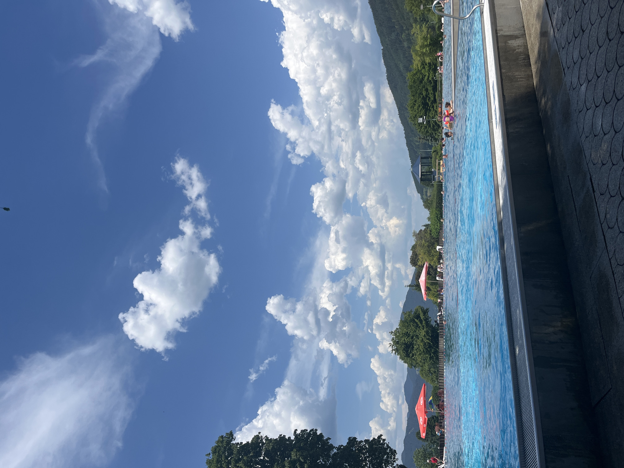 A sunny day at an outdoor swimming pool, likely the Freibad Oberammergau in Oberammergau, Germany. The scene is dominated by the bright blue water of the pool, with people swimming and relaxing. Red and pink umbrellas provide shade. Lush green trees and rolling hills form the backdrop under a vibrant blue sky filled with puffy white clouds. A paved walkway with a decorative pattern borders the pool, and a metallic grate runs along the pool's edge. The overall impression is one of leisure and recreation on a warm summer day.