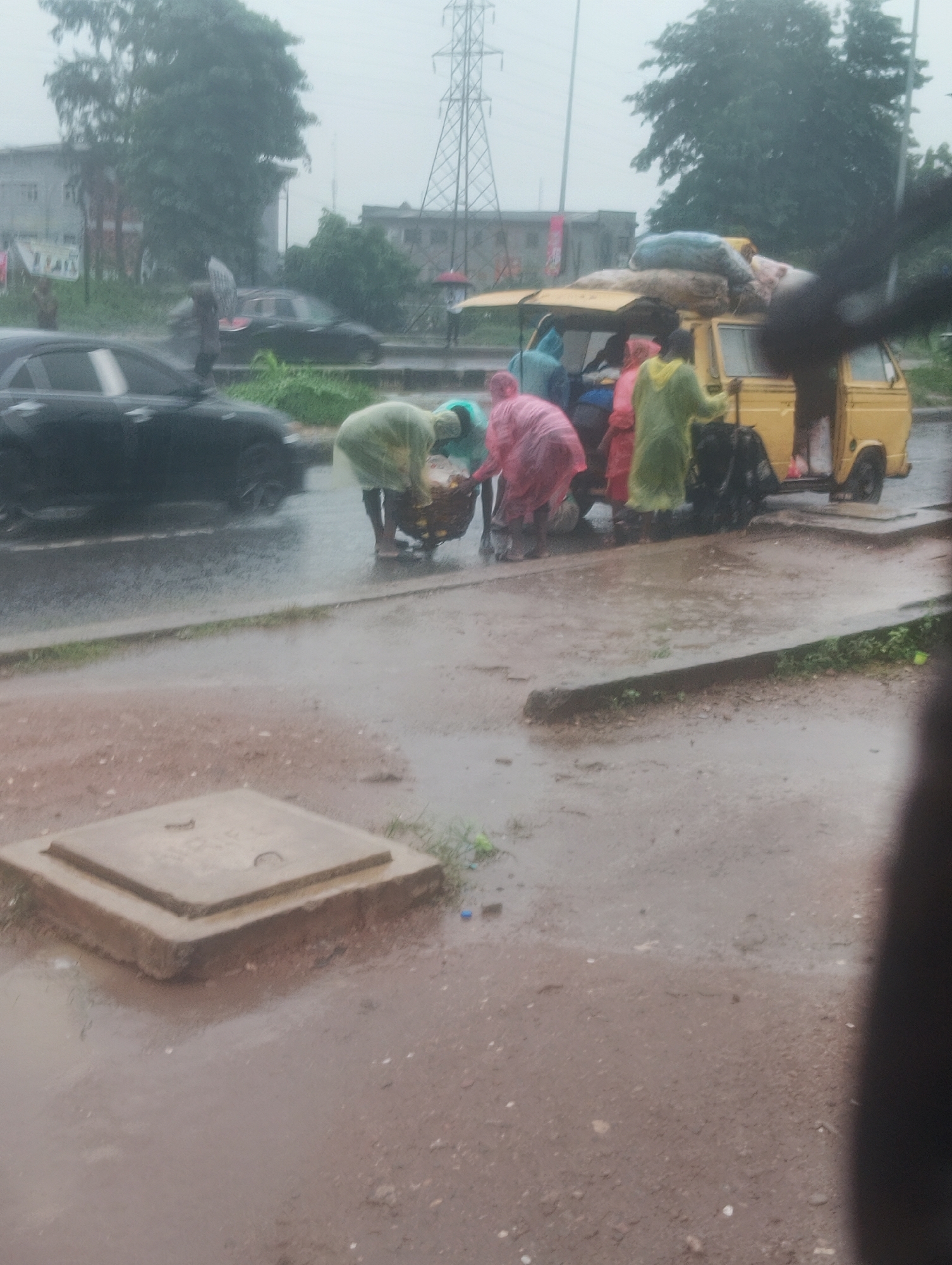 A scene unfolds in Ikeja, Nigeria, under a downpour. A yellow minivan, laden with luggage strapped to its roof, is parked on the side of a wet, asphalt road. Several individuals, clad in brightly colored raincoats – pink, green, and blue – are gathered around the van. Some appear to be loading or unloading cargo from the back of the van or from a basket on the ground. A black sedan is visible in the background, also on the road. Across the road, buildings and trees create a backdrop, with an electrical pylon standing tall in the distance. The ground is saturated with rainwater, creating puddles and reflecting the overcast sky. In the foreground, a square concrete manhole cover sits on the wet, reddish-brown earth. The overall impression is one of a busy street scene disrupted by heavy rain, highlighting the resilience of daily life in the face of inclement weather.