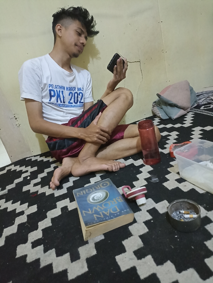 A young man is seated on a checkered rug, his right leg crossed over his left. He is wearing a white T-shirt with blue lettering that says "PELATIHAN KADER BANJ PKI 202" and red-and-black shorts. He is holding a black cell phone in his right hand and looking at it. The scene takes place in a room with a beige wall and a grey floor. To the right of the man are a red water bottle, a clear plastic container with a white substance, and a small black ashtray with two cigarette butts. A book titled "DAN BROWN ORIGIN" is lying in front of the man. This scene appears to have been captured in a room in Jatinangor, Indonesia, at the coordinates -6.934 latitude and 107.779 longitude.