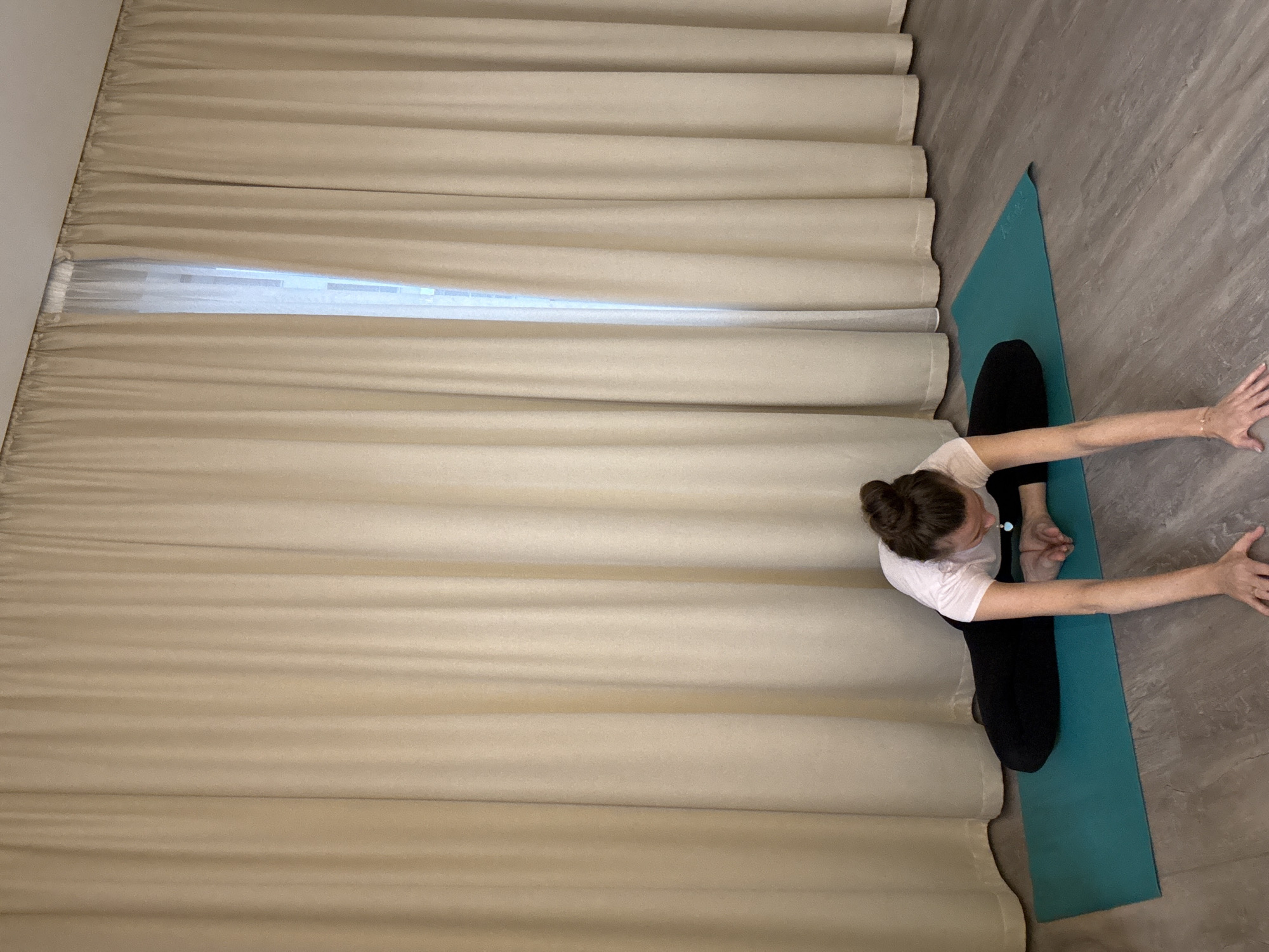A woman is performing a yoga pose on a teal mat in what appears to be a room with light beige curtains covering a window. The woman is wearing a white t-shirt and black leggings. Her hair is pulled back in a bun. She is leaning forward with her legs bent and the soles of her feet together, reaching her arms forward. The floor is a light-colored wood laminate. The scene is indoors and appears to be a yoga studio or a room in a home. The city of Dolgoprudny, Russia, is not depicted in this image.
