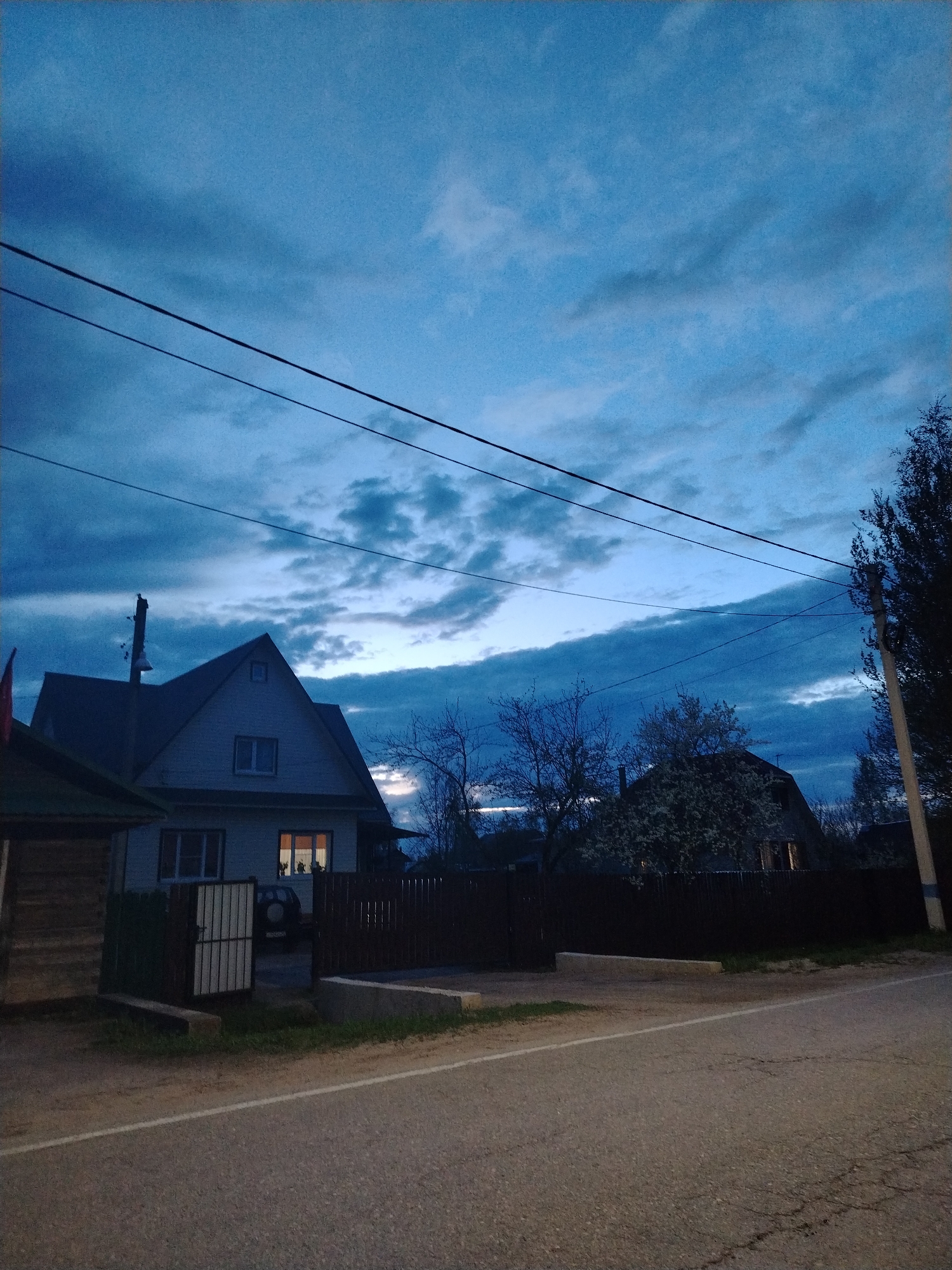 The image depicts a street scene in a residential area. A two-story house with a gable roof is visible on the left side of the frame, with light emanating from a window. A dark fence runs along the property line. The sky is overcast with dark blue and gray clouds. Overhead power lines stretch across the sky. A road runs along the bottom of the frame. To the right of the house, a tree with bare branches is visible, along with a second house partially obscured by a dark fence and foliage. A utility pole stands on the right edge of the frame. The scene appears to be at dusk or in the early evening.