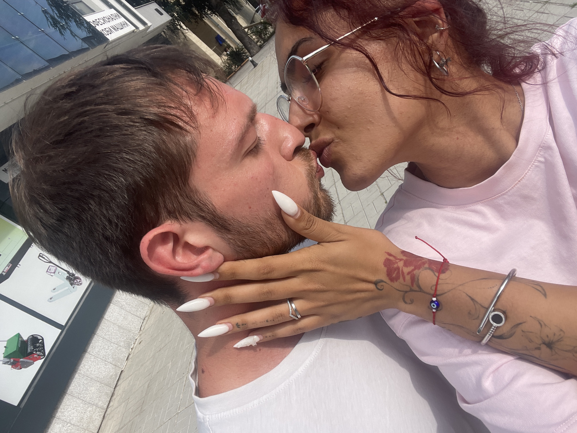 An overhead close-up depicts a male and female kissing outdoors. The male, positioned on the left, has brown hair, light skin, and a beard, wearing a white t-shirt. The female, on the right, has darker skin, dark red curly hair, and wears glasses, a pink t-shirt, and a thin silver necklace. Her right hand, adorned with long white stiletto nails, a silver two-band ring, and tattoos (a red floral design on the back of the hand/wrist, black abstract designs on the forearm, and small black symbols on the fingers), cups the male's cheek/neck. On her left arm, a silver nail-shaped bangle and a red string bracelet with a blue evil eye bead are visible, along with more black abstract tattoos on the forearm. Her right ear has multiple silver earrings. The background features a building with reflective windows showing trees and sky, a white sign with black Cyrillic text, and a display sign illustrating a green and red lawnmower-like machine and a white pallet jack. The ground consists of light-colored paving stones. The scene is brightly lit by daylight in the city of Unknown, Unknown.