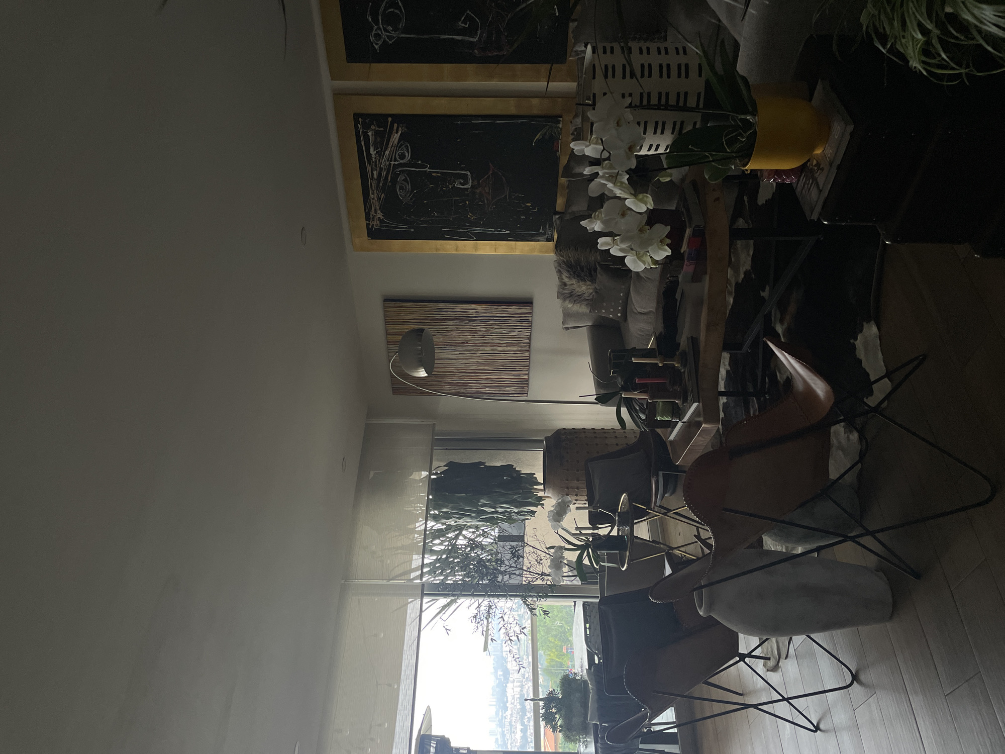 A brightly lit interior scene shows an apartment in Huixquilucan de Degollado, Mexico, filled with eclectic decor. The view through a large window features the cityscape, shrouded in a light mist. Inside, three butterfly chairs with leather seats and black metal frames are visible on a wood floor. In the background, abstract paintings hang on the wall above a contemporary floor lamp. A coffee table with white orchids and a yellow pot centerpiece sits in front of a grey couch. A tall plant is placed behind the window. The lighting is soft and diffused, creating a serene atmosphere. No people are visible.