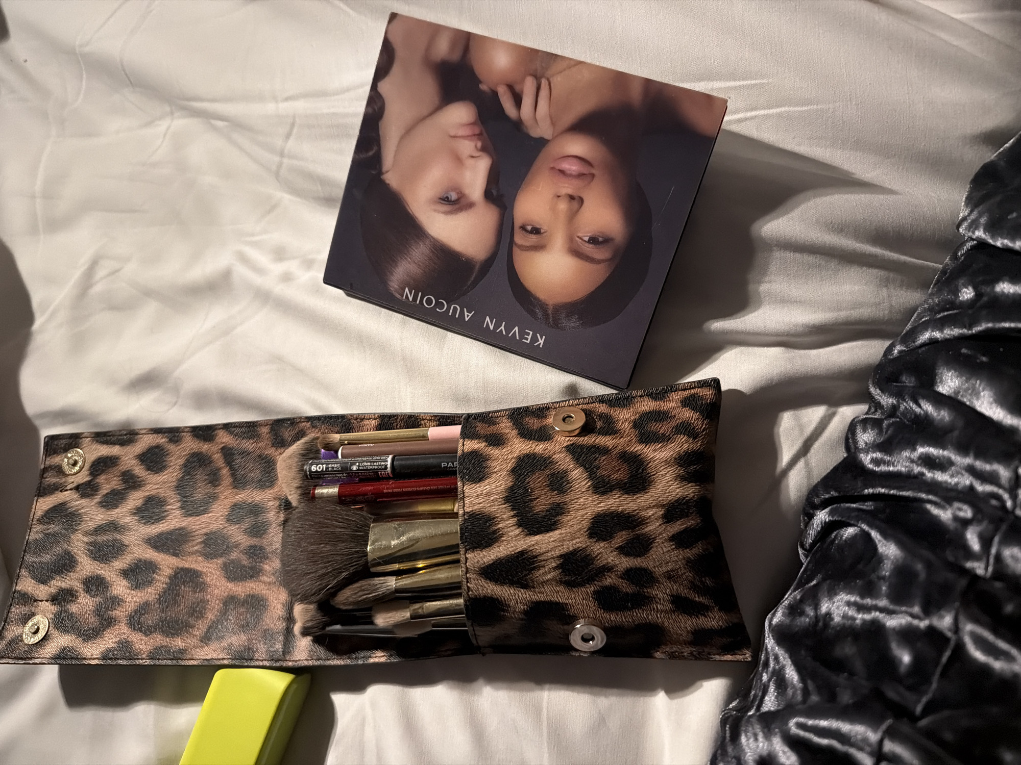 The image shows a makeup palette and makeup brushes on a white sheet or bedding, possibly in a bedroom. The makeup palette is a Kevyn Aucoin product featuring images of two women. A makeup brush case with a leopard print design is partially open, revealing various makeup brushes and pencils. A small, yellow object is visible in the lower-left corner. The presence of bedding and makeup items suggests a domestic, personal setting. There is no indication of the image being taken in Moscow, Russia.