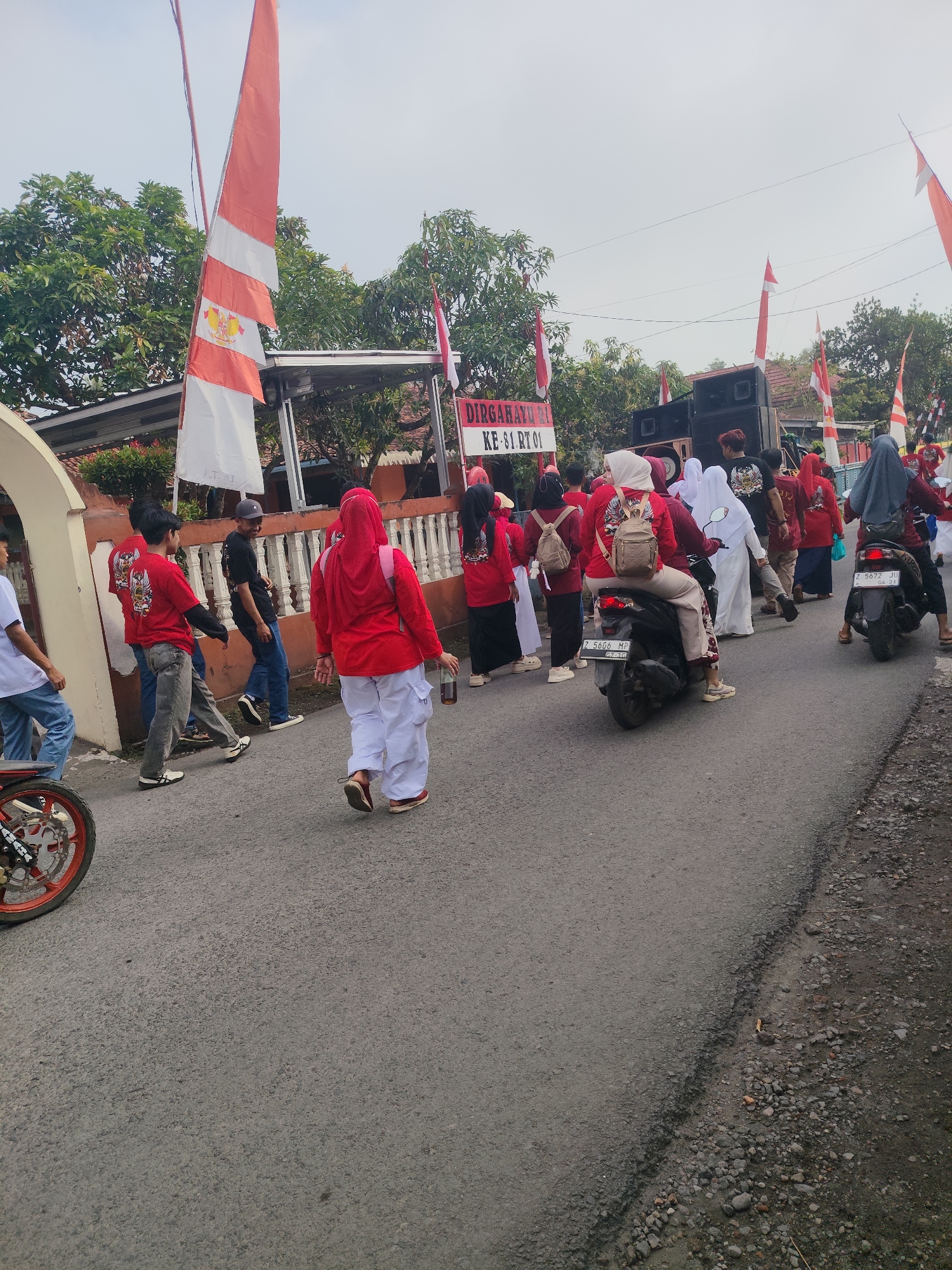 In Sukaratu, Indonesia, a group of people are walking and riding motorcycles down a paved road. Many are wearing red shirts and white pants, and some are wearing headscarves. There are red and white flags on poles lining the street, and a large speaker system is visible. The overall atmosphere appears to be festive, possibly a parade or celebration.