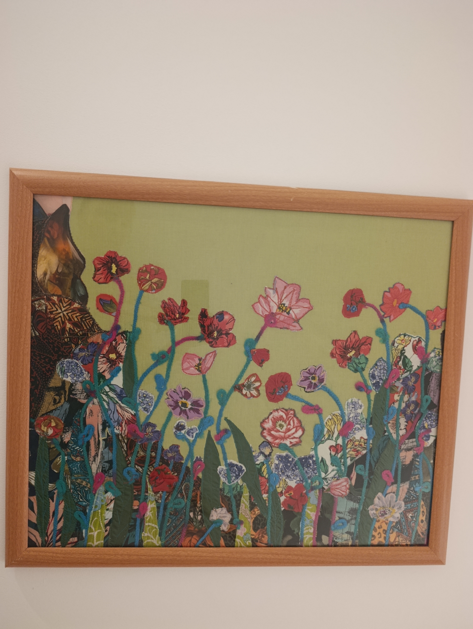 The image shows a framed piece of textile art, likely a collage and embroidery, depicting a field of flowers against a pale green background. The artwork is framed in light wood. To the left of the floral composition, a portion of a darker, patterned textile is visible, appearing to represent part of a larger image or a figure. The flowers are rendered in a variety of colors and textures, with some appearing to be embroidered with yarn. The background of the artwork has a subtle woven texture. The artwork is hung on a plain white wall. The scene is indoors. The specific location in Setúbal, Portugal cannot be determined from the image.