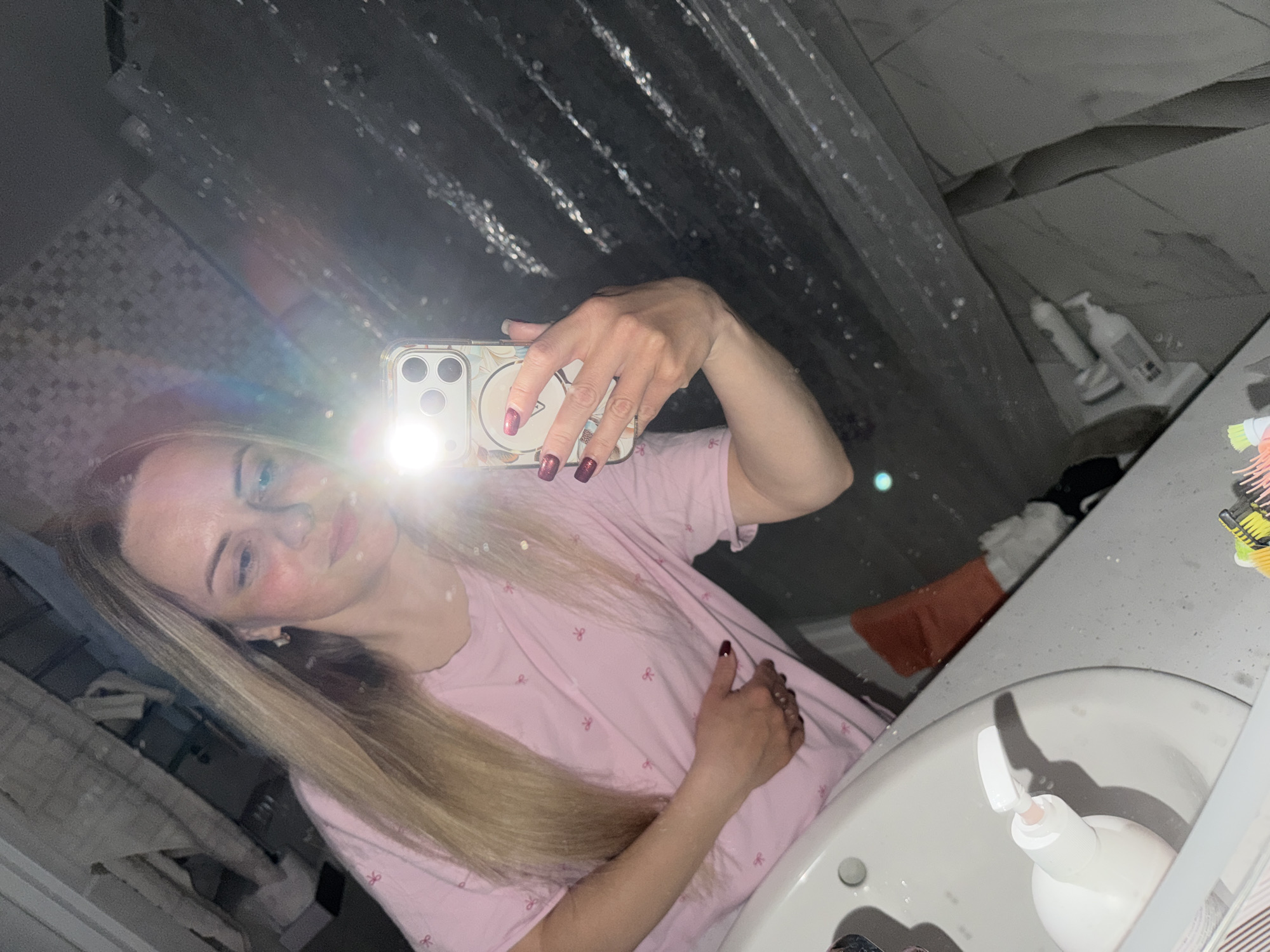 A woman is taking a selfie in a bathroom. She is holding a smartphone with a bright flash that obscures some of her face. She has long blonde hair and is wearing a pink shirt. The background includes a shower curtain, tiled walls, and a sink with a soap dispenser. There is no information to confirm the location in the city of Votkinsk, Russia.