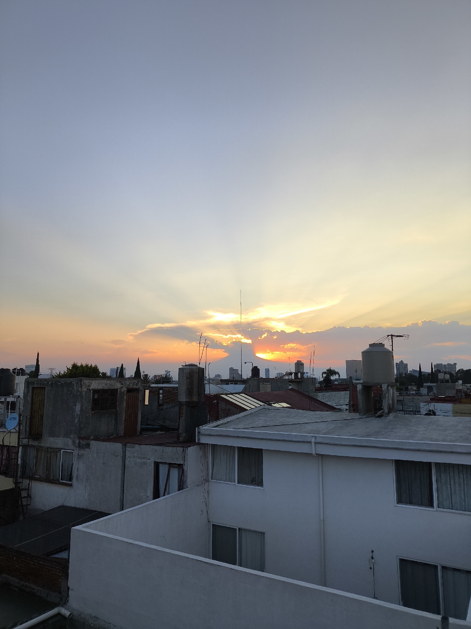 The image shows a cityscape at sunset, possibly taken from a rooftop in Puebla, Mexico. The sky is a gradient of light blue transitioning to orange and yellow near the horizon, with crepuscular rays emanating from behind clouds. The foreground consists of rooftops of buildings of varying heights and architectural styles. Some rooftops have water tanks, antennas, and other equipment. The buildings are mostly painted in shades of white and grey. Some windows are visible in the buildings. The background shows distant buildings, suggesting this is an urban area. There are no people visible in the image.
