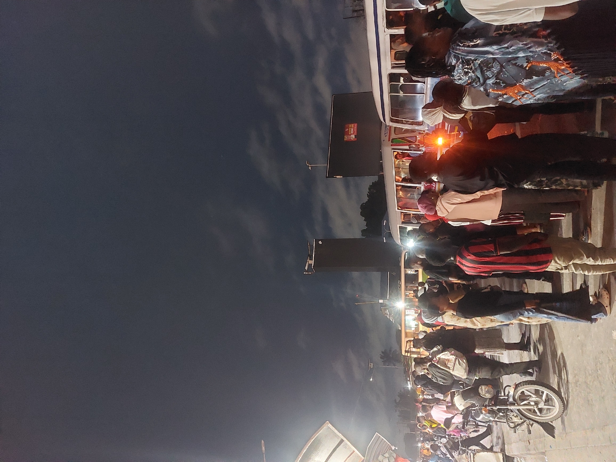At dusk in Dar es Salaam, Tanzania, people stand near a bus and parked motorcycles. A lit traffic light shines near a building. The sky is visible above the street.