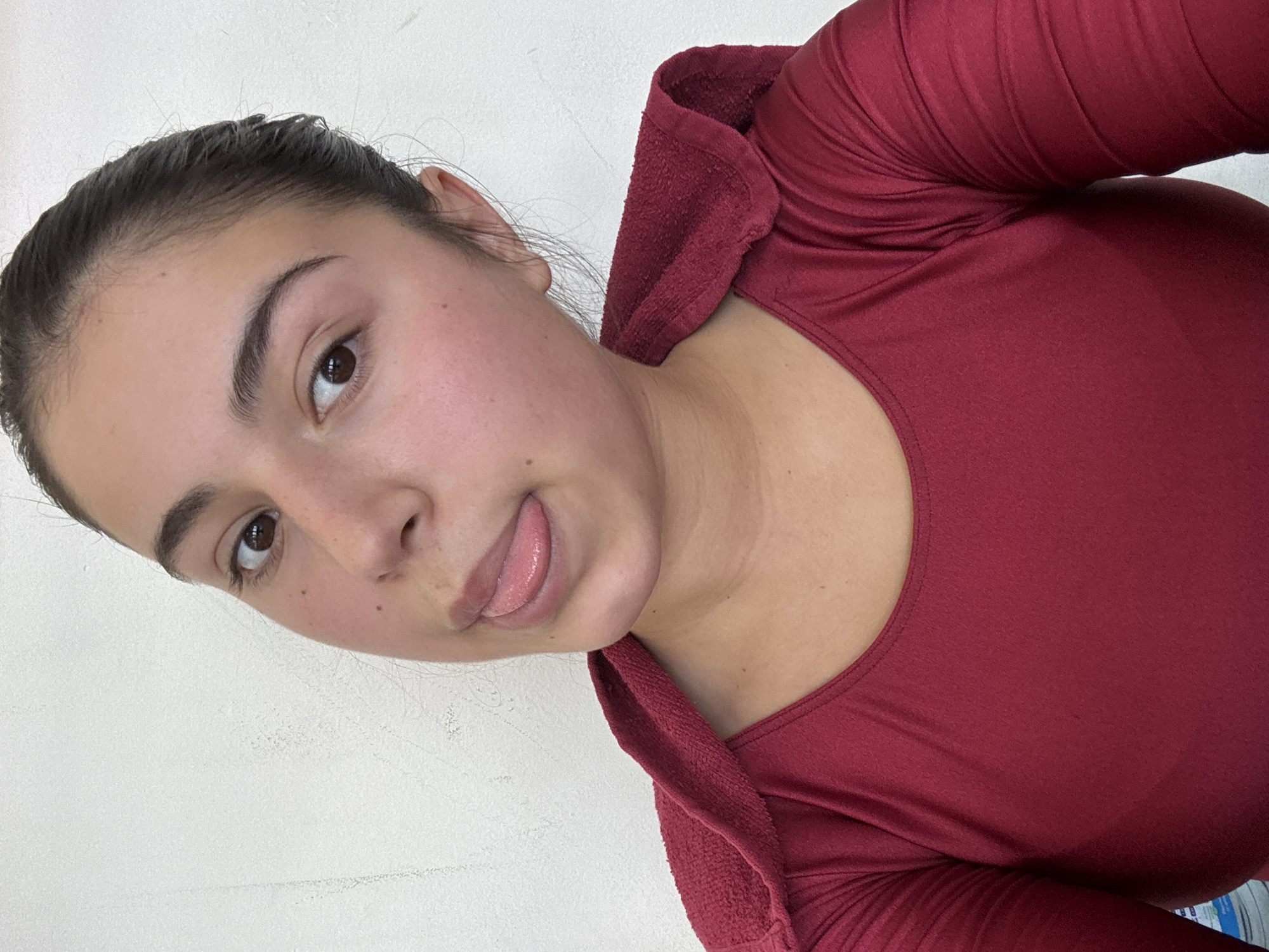 A close-up portrait features a young woman with dark hair pulled back from her face, possibly wet or slicked, set against a plain white background. Her head is tilted, giving the image a casual, dynamic feel, suggesting a selfie. She has dark, full eyebrows and brown eyes, looking directly at the camera. Her facial expression is playful, with her tongue sticking out slightly. Her skin appears clear with a few small moles visible on her face and neck. She is wearing a form-fitting, long-sleeved top in a deep red or maroon color. The main fabric of the garment is smooth, while a textured, slightly thicker fabric in the same color is visible around her neck and shoulder, suggesting a hood or a decorative collar detail. The lighting is bright and even, characteristic of an indoor setting. No text is visible in the image.