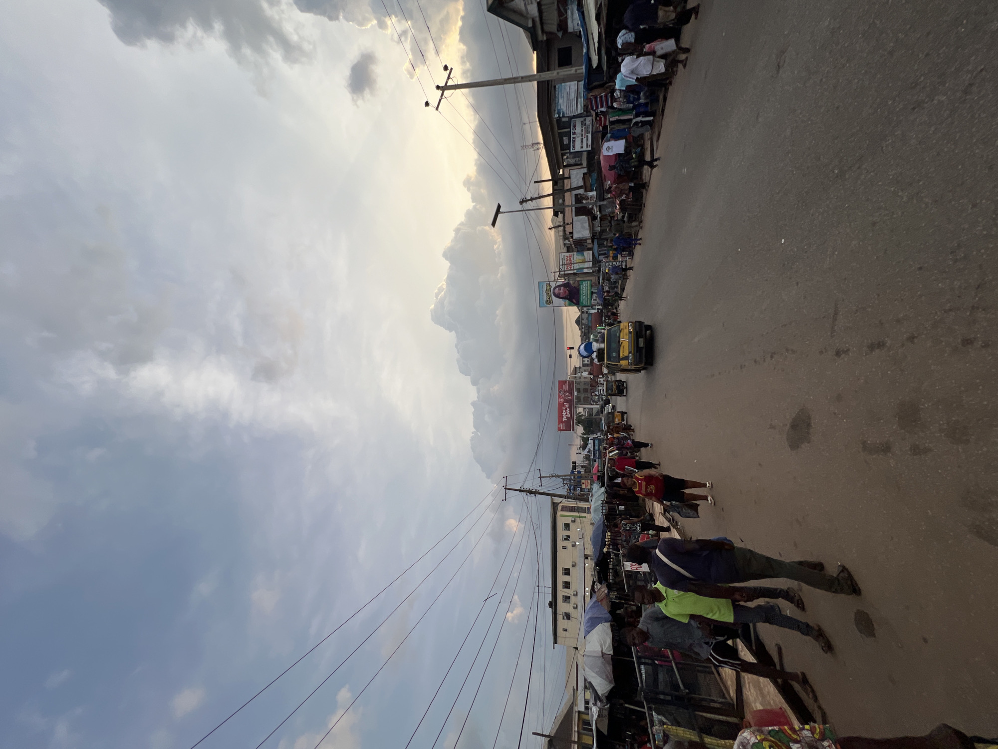 The image depicts a street scene in Nkpor, Nigeria, under a cloudy sky with a sunset glow. The street is paved and lined with shops and market stalls on both sides, covered by umbrellas and awnings. A mix of people are walking along the street and in front of the stalls. Some are carrying bags, while others are engaged in conversations. A yellow vehicle, possibly a taxi, is driving down the street. Utility poles with numerous wires are visible overhead, crisscrossing the sky. Billboards are also present on the buildings. The overall impression is that of a busy marketplace in a Nigerian urban setting during the late afternoon or early evening.