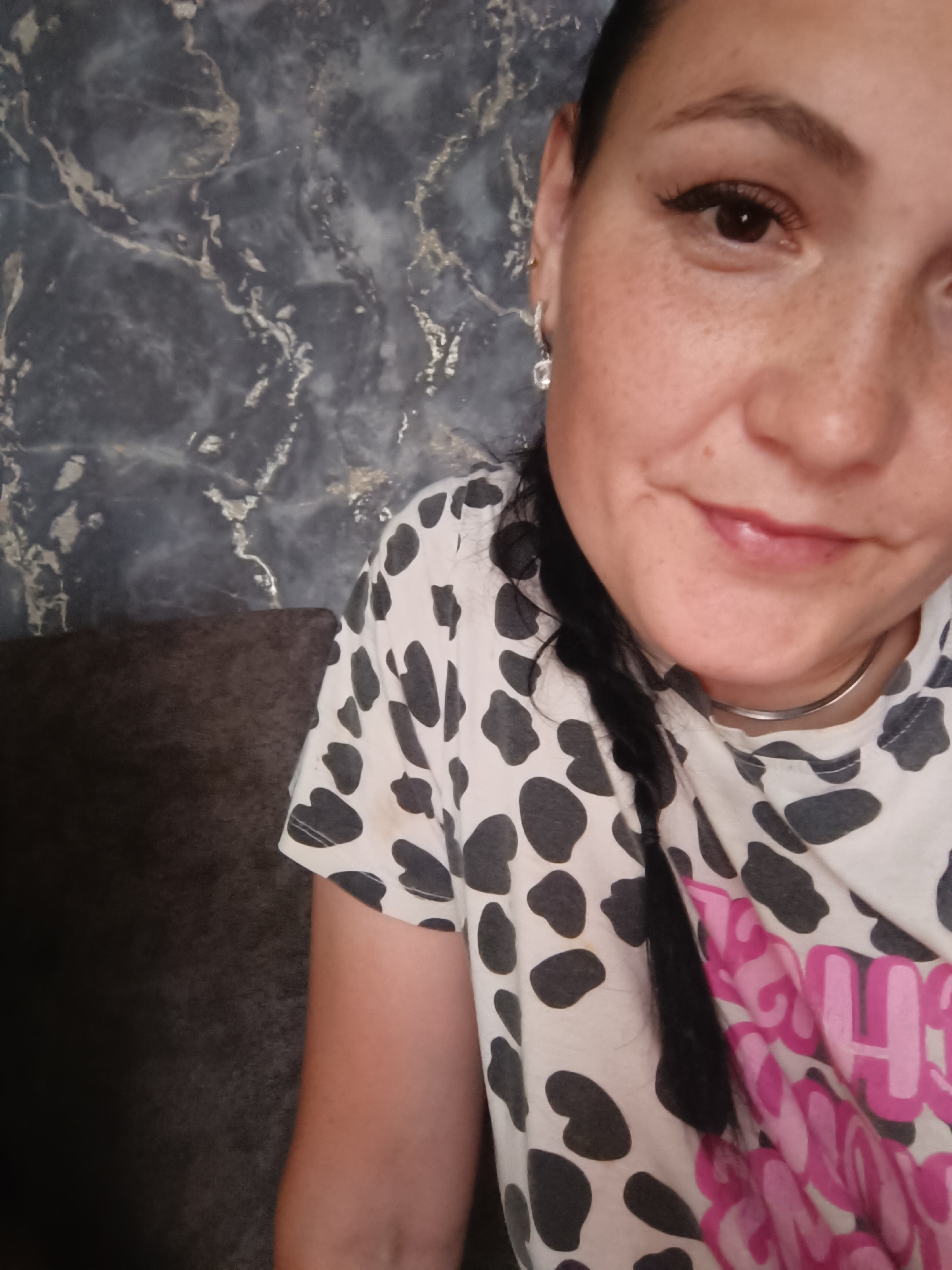 A young woman is captured in a close-up, selfie-style portrait. She has dark hair styled in a braid that falls over her shoulder. Her face is turned slightly towards the camera, and she is smiling. She is wearing a white t-shirt with a black cow-print pattern, and a pink graphic design is visible on the lower part of the shirt. She is also wearing a thin silver necklace and a dangly earring with a clear stone. The background appears to be a wall with a dark marble pattern, and a dark-colored couch is visible on the left side of the image. No specific location in the city of Karsha, Russia is identifiable in this image.