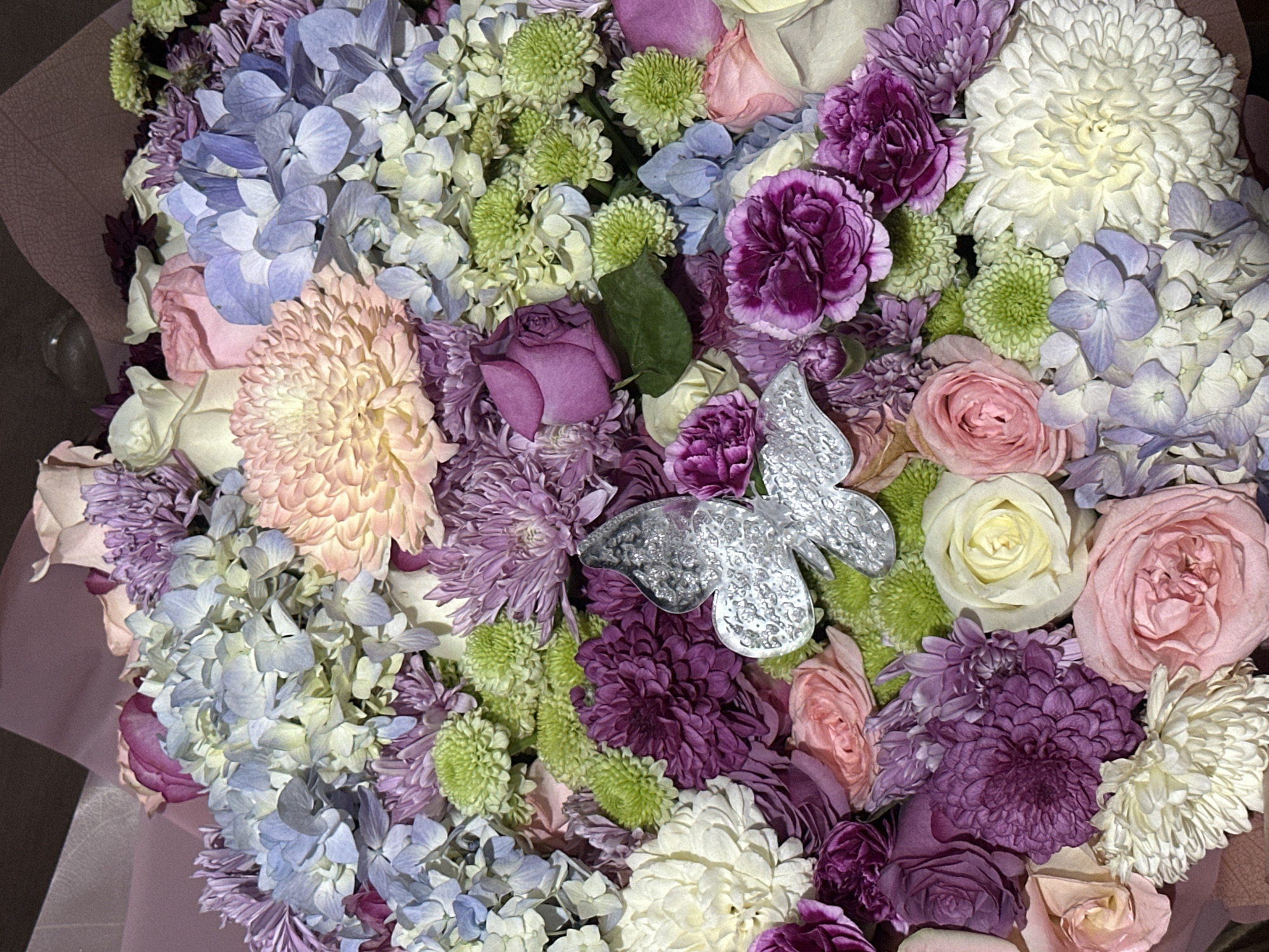 A close-up shot shows a bouquet of various flowers, including hydrangeas in shades of blue and light purple, pink and white roses, green spider mums, purple carnations, and lavender chrysanthemums. A silver butterfly ornament is placed among the purple flowers. The bouquet is wrapped in pink paper. The scene's location is Unknown, Unknown.