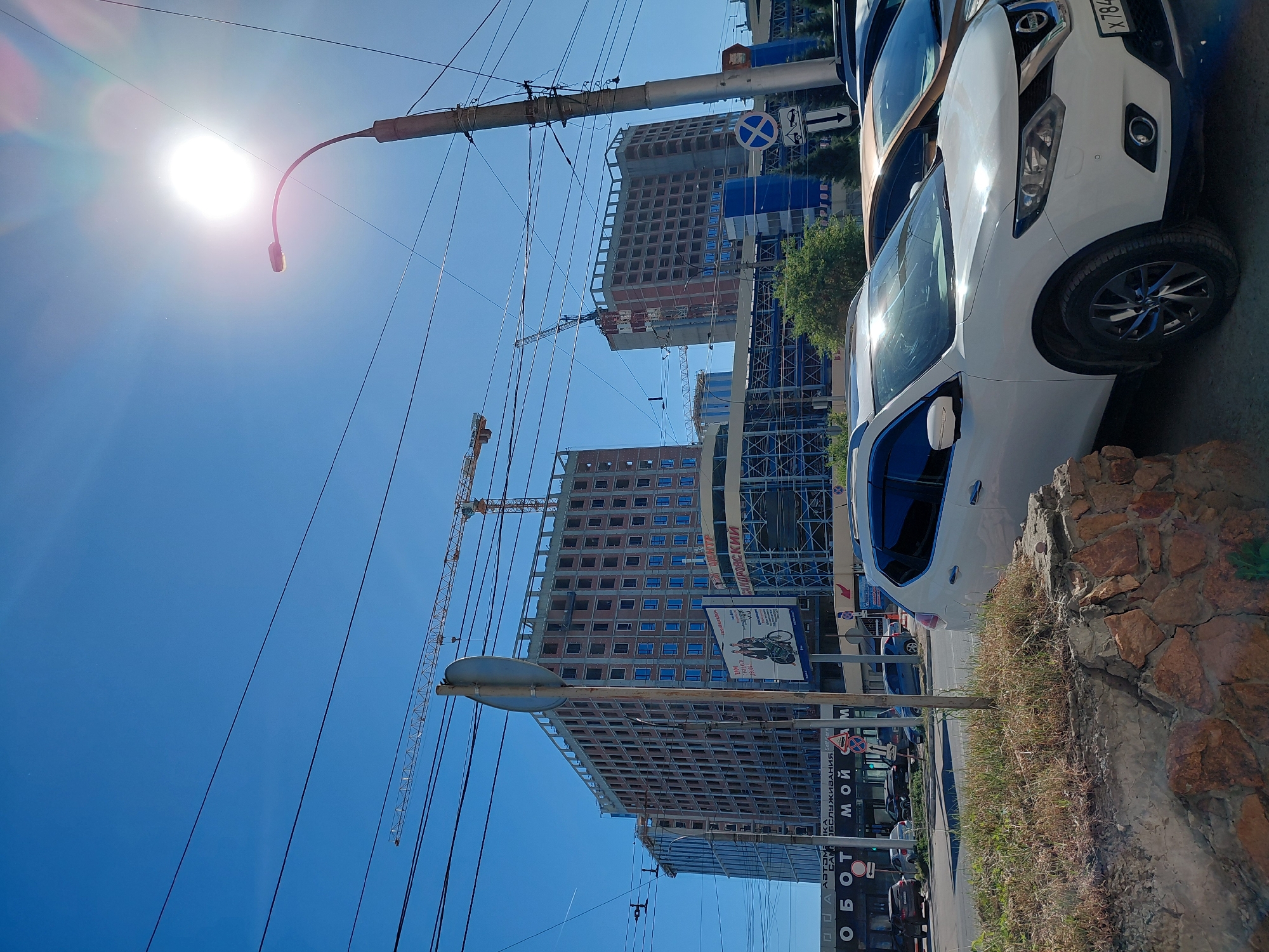 A bright, sunny day in a city with clear blue skies dominates the scene. In the foreground, a white SUV is parked, its windshield reflecting the sun. Behind it, other cars are visible. The middle ground features a street with several vehicles driving. A large, multi-story building under construction with exposed brickwork and blue-tinted windows is prominently featured. Construction cranes are visible above the building. Further back, another similar building, also under construction, is present. A street lamp with a curved arm stands on the right side of the image. Power lines crisscross the sky. In the background, there are more buildings and trees. Signage in Russian is visible on some of the buildings, including "ОБОТ МОЙ" and "ТОРГОВЫЙ ЦЕНТР". The overall impression is a bustling urban environment with ongoing development.