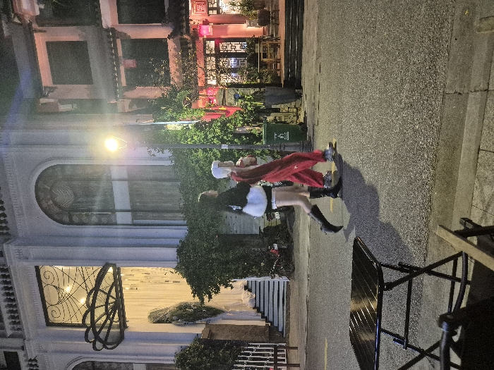 This nighttime image shows a street scene in Thuận Hóa Ward, Vietnam. Two individuals are walking from left to right across an asphalt road surface illuminated by a bright streetlight. The person on the left wears a black long-sleeved top, white shorts, and black knee-high boots with heels. The person on the right wears a white cap, a light-colored top, red pants, and light-colored athletic shoes. Shadows are cast behind them due to the light source.
To the left, a multi-story building with a light-colored facade features decorative architectural elements, large arched windows, and a dark, cloud-shaped canopy above an entrance. White fabric or light strips descend from one window. White stairs lead up to an entry, with a bicycle or scooter partially visible parked nearby. Green foliage grows along the building's base.
To the right, a darker, multi-story building with traditional East Asian architectural influences features square windows and red lanterns at its lower level. Wooden tables and benches are visible at its entrance. Green vines climb the building's facade and the streetlight pole. A green cylindrical trash can stands near the pole on the paved sidewalk. In the lower left foreground, a dark, foldable outdoor table with horizontal slats is partially visible on the sidewalk, with a portion of a wooden chair armrest in the extreme bottom left.