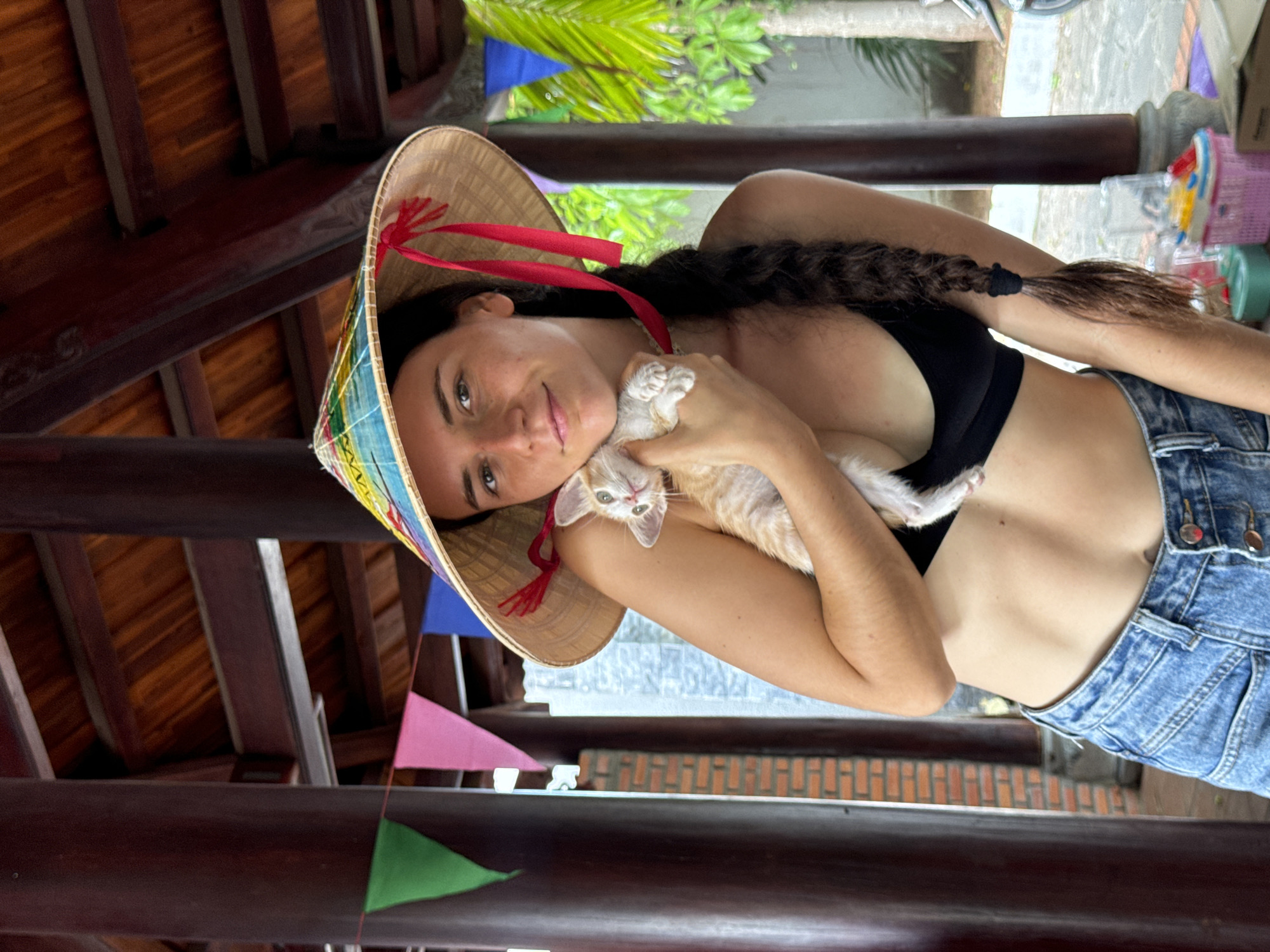 A young woman with dark hair braided down her back wears a conical hat decorated with colorful patterns and red ribbons. She is dressed in a black sports bra and blue denim shorts, revealing her toned midriff. The woman is holding a small, light-colored kitten close to her chest. They are positioned under a wooden-roofed structure with dark brown pillars. In the background, colorful triangular flags are strung across, and palm trees are visible. The setting appears to be an outdoor area in Mũi Né, Vietnam, possibly a resort or a market stall given the presence of what looks like decorative items and baskets on a table in the lower right corner.