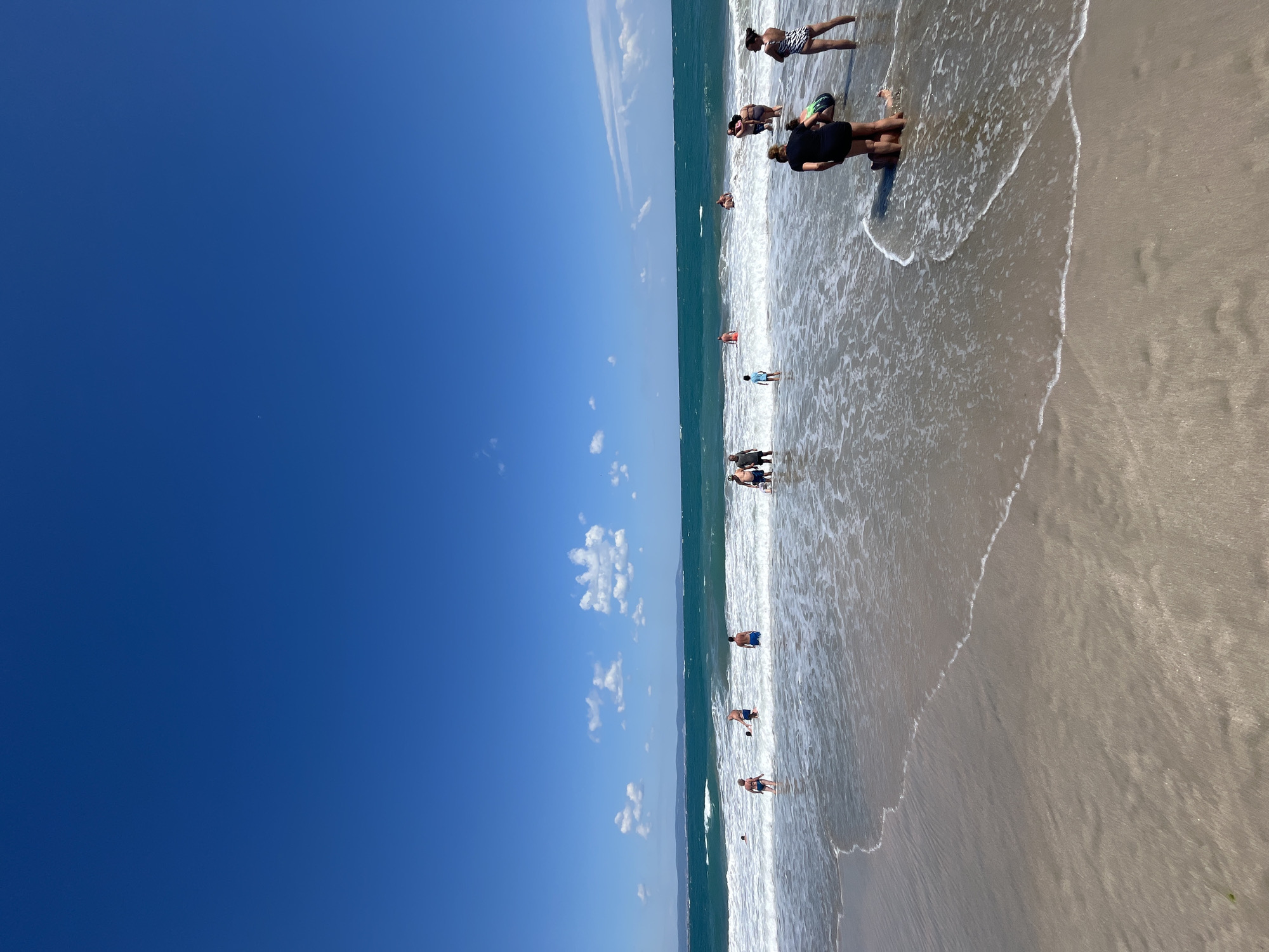 This is a full-frame, eye-level, outdoor, vertical shot of a beach in Pomorie, Bulgaria, on a bright, sunny day. The scene captures people enjoying the water as waves break on the shore. The sky is a vibrant, clear blue with a few scattered white clouds. The ocean is a deep turquoise color, with white foamy waves rolling in towards the sandy beach. In the middle ground, a group of people are wading in the shallow water, some with swimsuits on. Some are standing, while others are partially submerged. In the foreground, the wet sand is visible with the gentle lapping of waves. The overall impression is a lively and refreshing summer scene at the seaside.