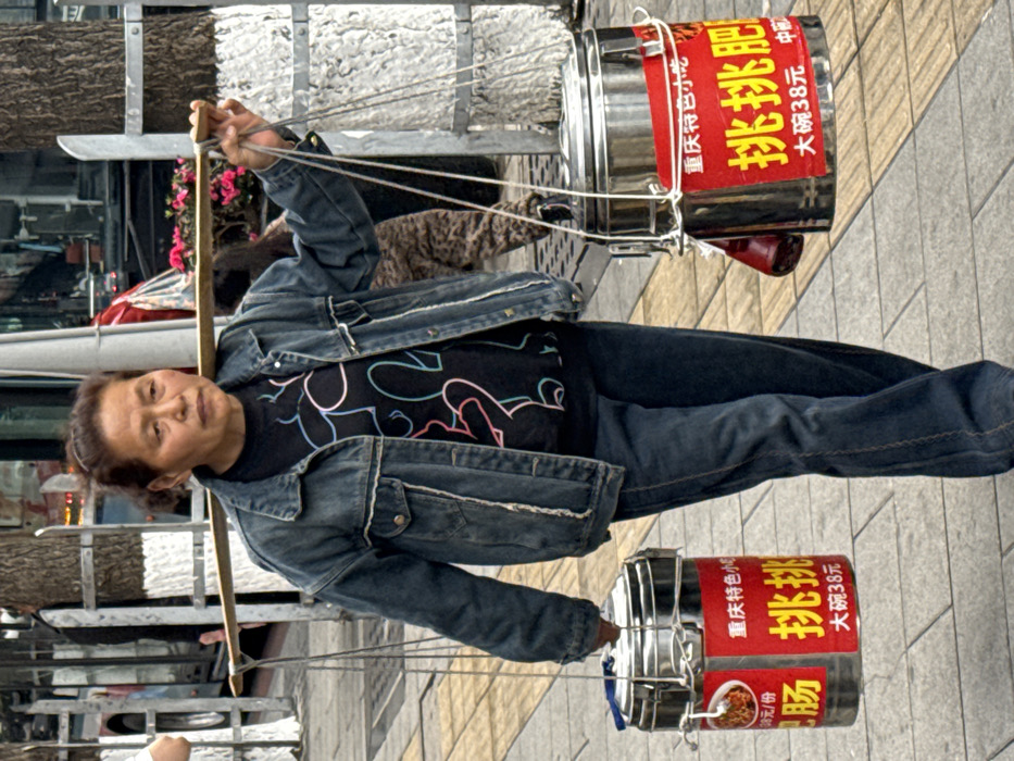 An older person is carrying two stacks of metal containers using a wooden pole across their shoulder. The containers have red labels with yellow and white Chinese text. One label reads "Chongqing Specialty Small Intestine, Pickled肥 (féi), Large Bowl 38 Yuan." The person wears a denim jacket and dark pants. The scene appears to be set on a paved street.