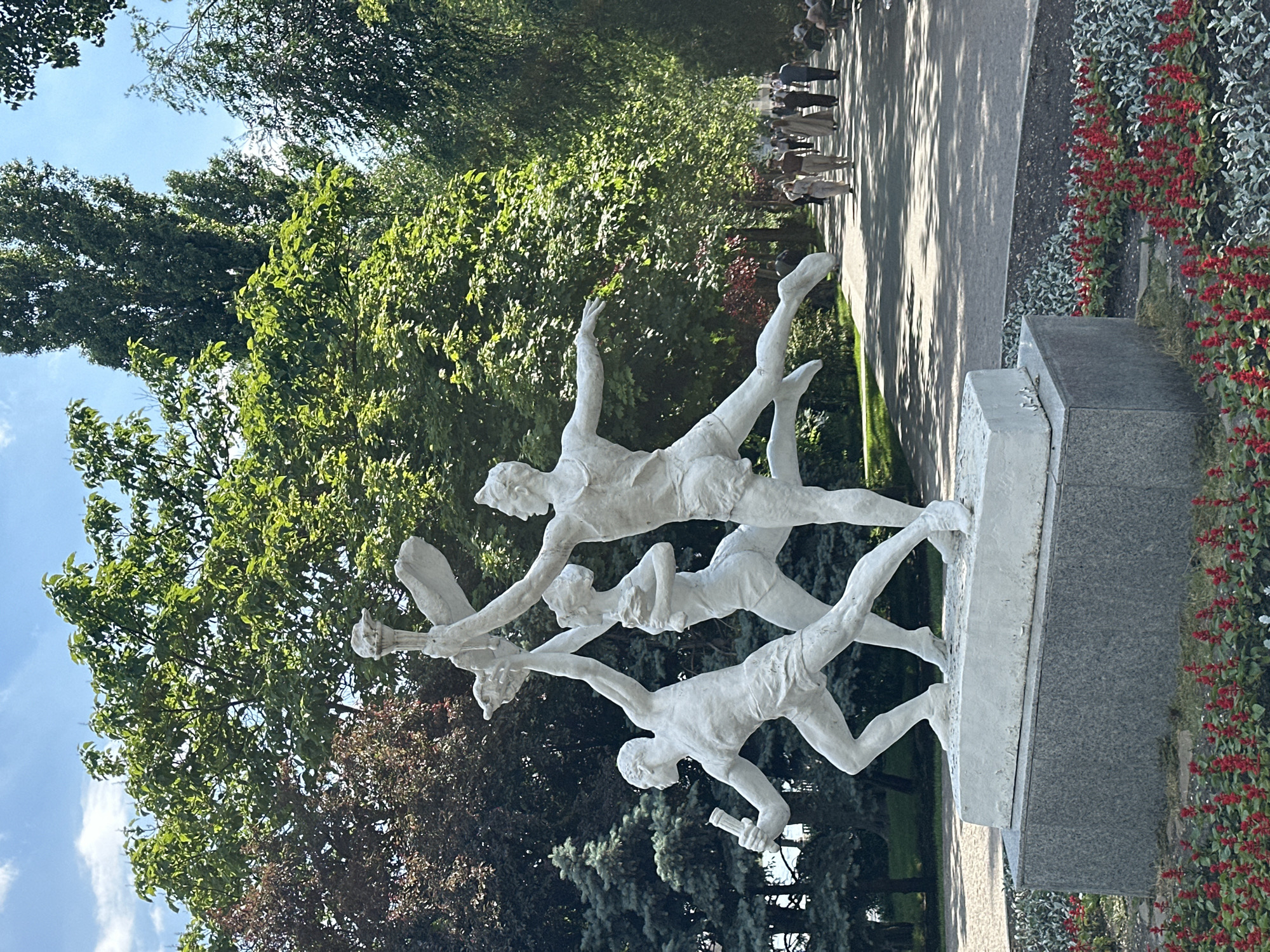The image displays a bright, sunlit park scene in Rostov-on-Don, Russia, centered on a white, multi-figure sculpture. The sculpture, positioned slightly left of the frame's center, depicts three athletic individuals in dynamic motion, suggestive of a relay or celebratory run. The leftmost figure is a male, striding forward, holding a torch-like object aloft in his right hand. The central figure is a female, running with both arms raised, clutching a wreath-like object. The rightmost figure, a male, is captured in a mid-leap pose, holding a stylized flag or banner overhead in his left hand. The figures are sculpted in a monochromatic white finish, resting upon a rectangular white plinth, which in turn sits on a dark grey, textured pedestal, likely granite.
In the immediate foreground, surrounding the pedestal, a flower bed exhibits clusters of vibrant red flowering plants (possibly Salvia) intermingled with silvery-leaved plants (possibly Dusty Miller). A paved, light grey pedestrian pathway occupies the right side of the midground, curving into the distance. Approximately eight to ten individuals are discernible walking along this path, dressed in casual clothing.
The background features a dense canopy of trees. To the left, there are dark green coniferous trees and reddish-brown deciduous trees. To the right, tall, lush green deciduous trees line the pathway, creating an archway effect and casting dappled shadows on the ground. The sky above is clear blue with sparse white clouds, indicating bright daylight conditions.