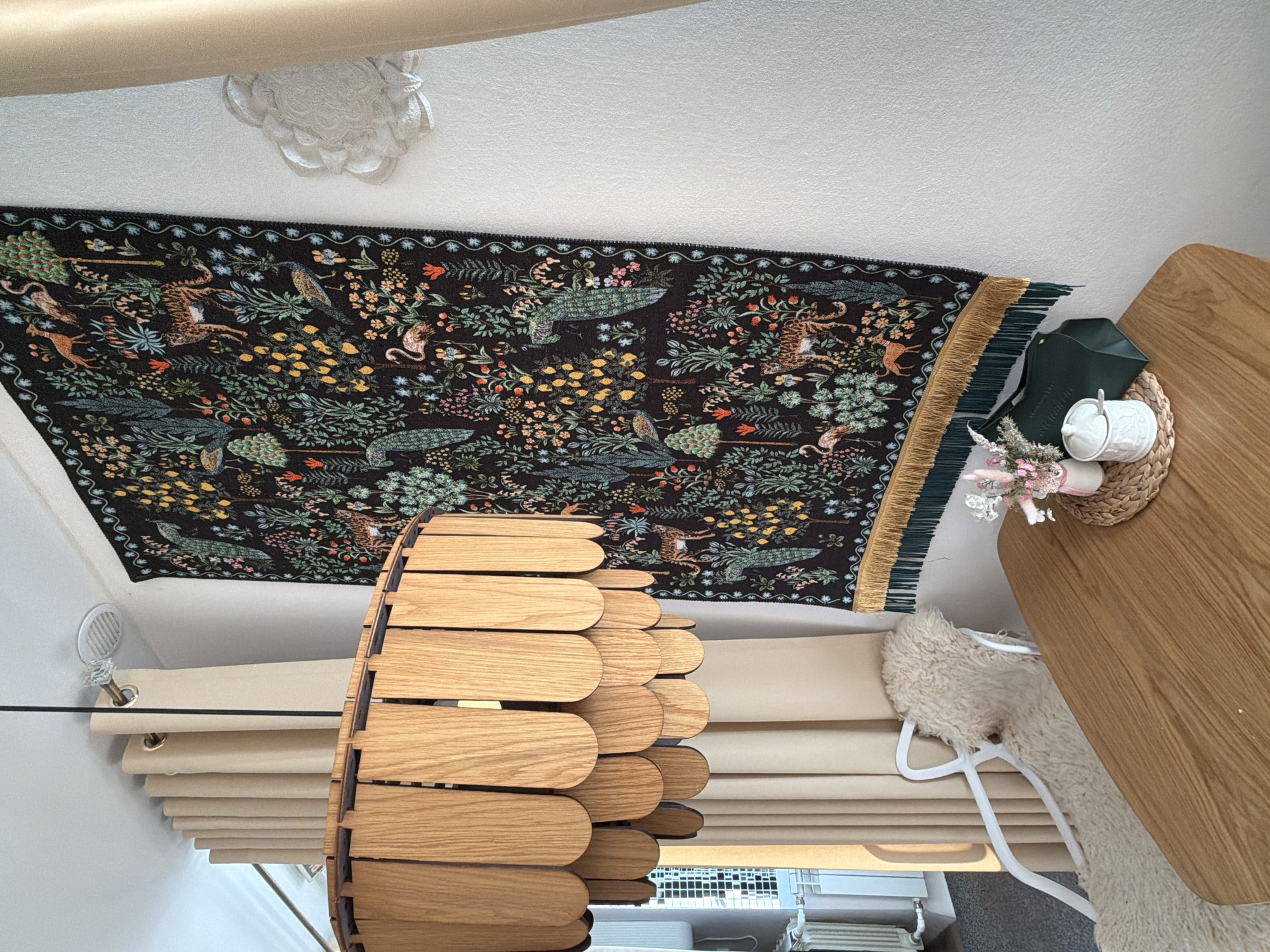 The image presents an interior scene, likely indoors. Dominating the foreground is a wooden, cylindrical light fixture with a slatted design. Behind the light fixture, a set of beige window blinds is visible. Above this, a patterned rug or tapestry is hung on the wall, featuring a dark background with detailed botanical and animal illustrations. To the right, a wooden table is visible, with a woven placemat on top holding a small white container with a spoon and a dark green pouch. A floral arrangement is also present on the placemat. A white, fur-like object is partially visible, possibly a seat cushion. The background suggests a modern or decorated interior, with a white ceiling adorned with a decorative molding. The scene appears to be a room setting, possibly a dining area or living space. The location in Votkinsk, Russia cannot be confirmed or disproven.