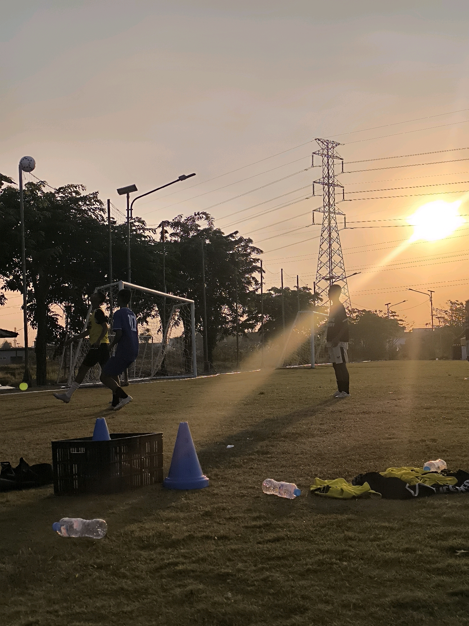 At sunset on a grassy field, two young males are playing soccer. The individual in the yellow jersey is leaping to head a ball. Another young male, dressed in a blue jersey with the number 10, is standing near the goal. The scene is lit by the setting sun, casting long shadows across the field. In the background, trees, streetlights, and a tall power pylon are visible. Scattered on the grass are sports equipment, including cones, a black crate, and a few bottles of water. The presence of the power pylon and the general urban landscape suggest this location is likely within the city of Surabaya, Indonesia.