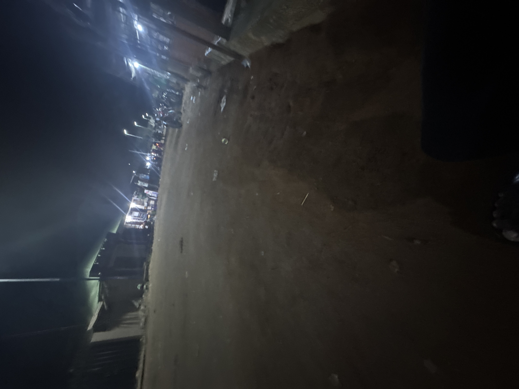 The image shows a dark, unpaved road in Ihiagwa, Nigeria, at night. Streetlights illuminate a row of buildings and shops in the distance. Parked motorcycles are visible near the shops. A dark, indistinct mass and human toes are in the foreground on the right side of the frame. Debris is scattered on the road surface.