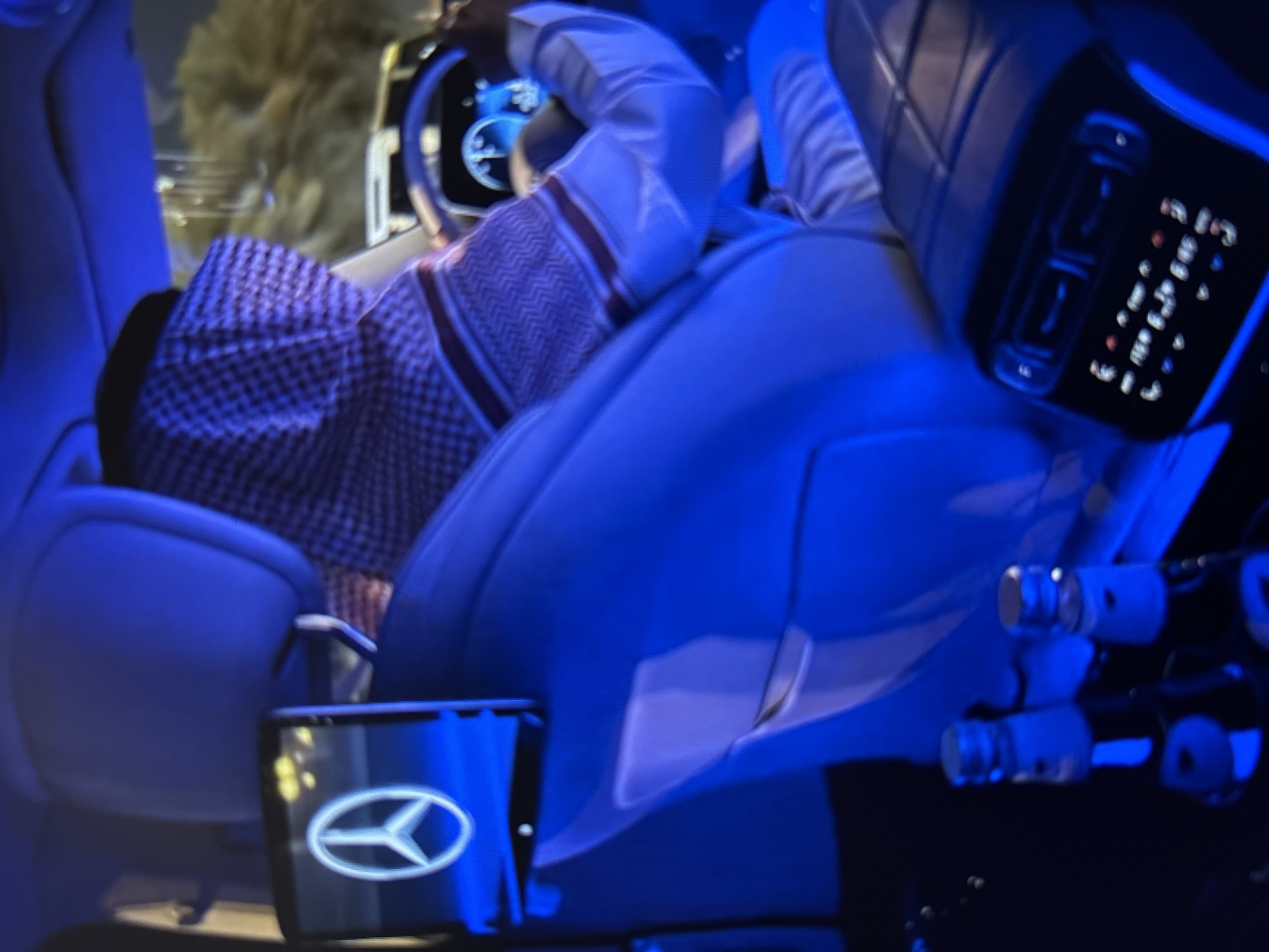 An interior view of a luxury vehicle, illuminated by blue ambient lighting. A person, dressed in traditional Saudi attire with a red and white checkered ghutra, is seated in the driver's position, holding the steering wheel. A tablet displaying the Mercedes-Benz logo is mounted on the back of the driver's seat. In the foreground, two bottles are visible, possibly beverages. The vehicle appears to be in motion, as indicated by the blurred background. The scene is set at night, with lights from the surroundings visible through the windows. The car is likely located in an urban environment, though no specific landmarks in Burgas, Bulgaria, are discernible in the image.