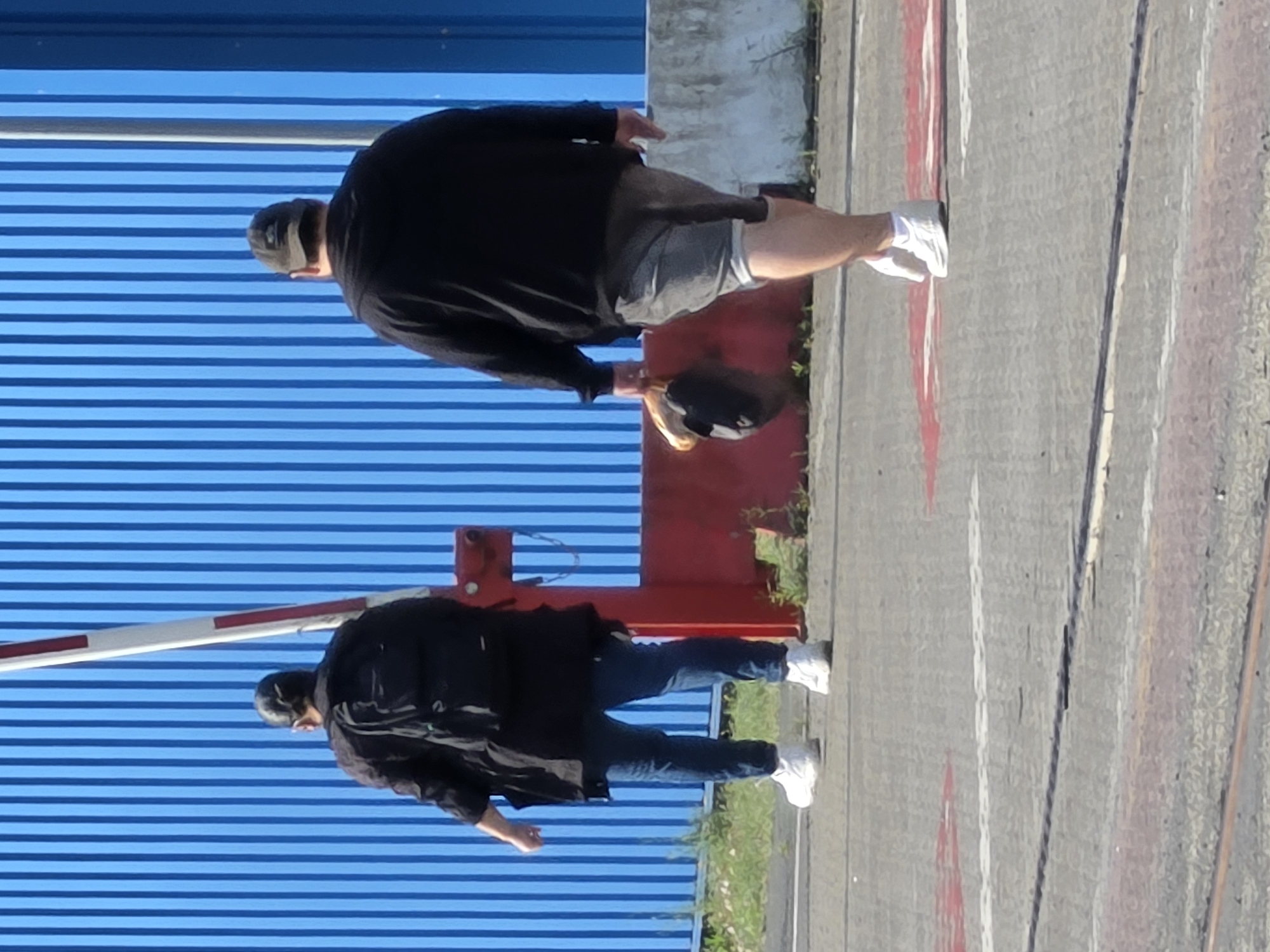 The image shows a man and a woman walking away from the camera on a paved surface, likely a street or industrial area. Both are dressed in casual clothing and walking towards a large blue corrugated metal structure. The man on the right is wearing a dark hooded sweatshirt, grey shorts, and white sneakers. He is carrying a dark baseball cap with a tan brim in his right hand. The woman on the left is wearing a dark jacket, blue jeans, and white sneakers, and has a black backpack on. Behind them, a red and white barrier gate is partially visible, and further back, a bright blue corrugated metal wall dominates the background. The ground is marked with white and red lines, suggesting a designated pathway or road. The lighting suggests it is daytime, with shadows cast on the ground. The scene appears to be in an urban or industrial setting. The query about Frankfurt, Germany, cannot be visually confirmed from the image content.