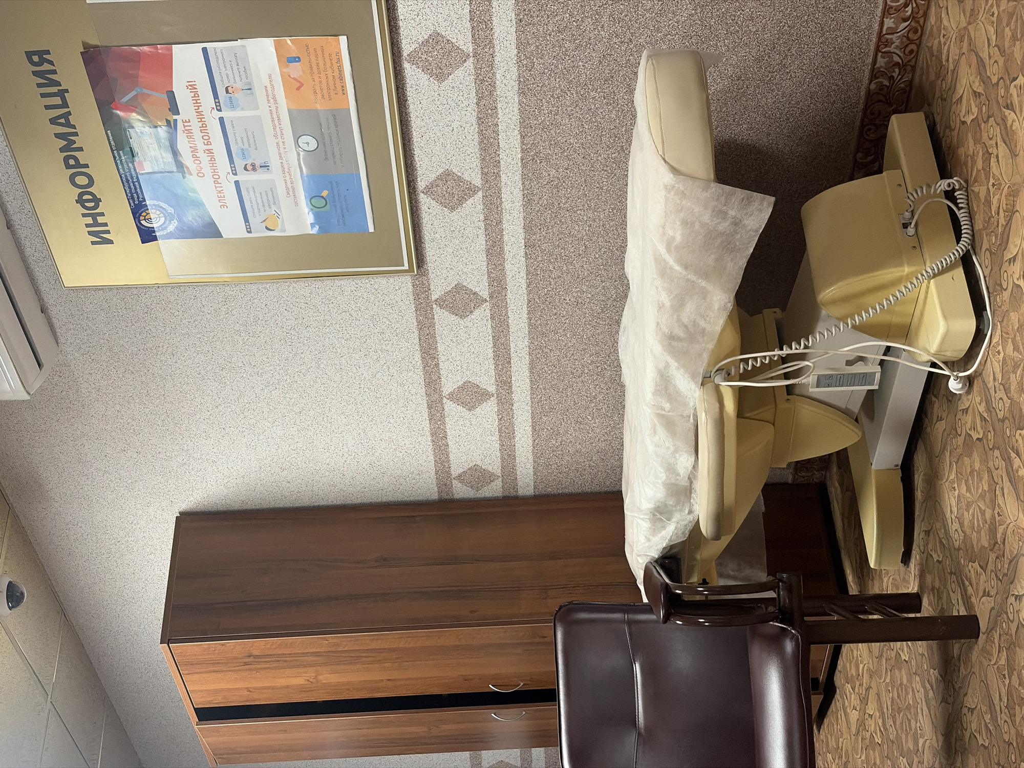 This image captures a functional room, likely a medical examination room or a clinic waiting area in Saint Petersburg, Russia, as indicated by the text. No people are present, and no specific activity is taking place.
The prominent feature is a beige medical chair or examination table, covered with a white disposable sheet, suggesting readiness for a patient. Wires extend from the chair, connecting to a small display or control unit near its base. To the left, a dark brown wooden cabinet with two drawers is visible. In the foreground, a dark brown leather chair with wooden legs faces away from the viewer.
The room's walls are covered in light-colored wallpaper featuring vertical stripes with repeating diamond patterns. The floor has an intricate, brownish, floral-like pattern. Partially visible on the far left is what appears to be an air conditioning unit.
A large, framed informational poster dominates the upper left portion of the wall. The heading reads "ИНФОРМАЦИЯ" (INFORMATION). Below this, it identifies the "Санкт-Петербургское региональное отделение Фонда социального страхования Российской Федерации" (Saint Petersburg regional branch of the Social Insurance Fund of the Russian Federation). The main message on the poster is "ОФОРМЛЯЙТЕ ЭЛЕКТРОННЫЙ БОЛЬНИЧНЫЙ!" (APPLY FOR AN ELECTRONIC SICK LEAVE!). The poster details the process: obtaining an electronic sick leave from a doctor, informing the employer, and receiving benefits. It also highlights benefits like medical confidentiality ("Сведения о вашем диагнозе, обследовании и лечении составляют врачебную тайну и не станут известны работодателю"), efficient record-keeping ("Контроль за правильностью расчетов и архив ваших электронных больничных в Личном кабинете. www.cabinets.fss.ru"), and time-saving ("Экономия времени. Ненужно стоять в очередях и собирать подписи врачей"). The setting appears to be indoors during daytime, under artificial lighting.