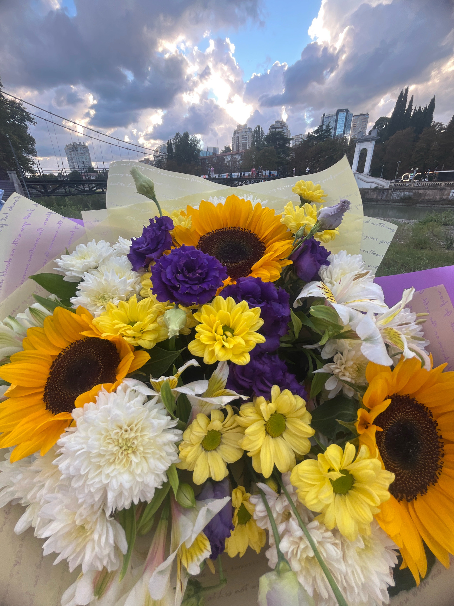 A bouquet of sunflowers, yellow chrysanthemums, white chrysanthemums, purple lisianthus, and white lilies is presented in the foreground. The bouquet is wrapped in paper with handwritten text and a purple ribbon. In the background, a bridge with a unique, cylindrical building atop one of its supports spans across a body of water. Further in the distance, a city skyline with modern buildings and cypress trees is visible. The sky is cloudy with sunlight breaking through. This scene is located in Sochi, Russia, near the Primorsky Bridge. There are no people visible in the image.