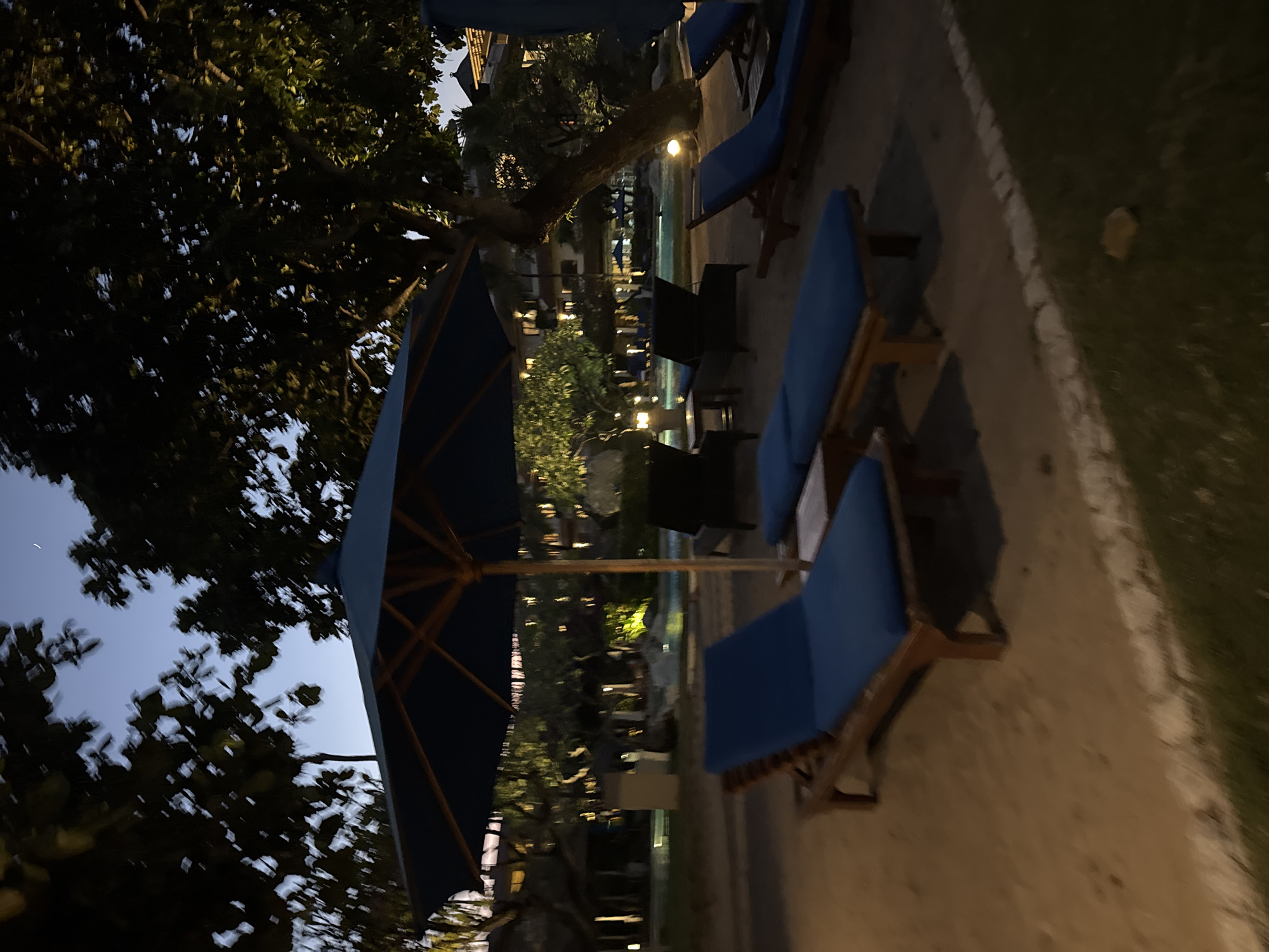 The image depicts a resort setting in Kuta Selatan, Indonesia, during dusk. In the foreground, two blue lounge chairs with wooden frames are positioned on a sandy area, with another lounge chair partially visible to the right. A large blue umbrella stands prominently in the center, casting a shadow. Beyond the lounge chairs, a swimming pool is visible with surrounding trees and buildings, some of which are illuminated by lights. The sky is a deep blue with a faint crescent moon. The overall atmosphere is tranquil, suggesting a relaxing evening at a tropical resort.