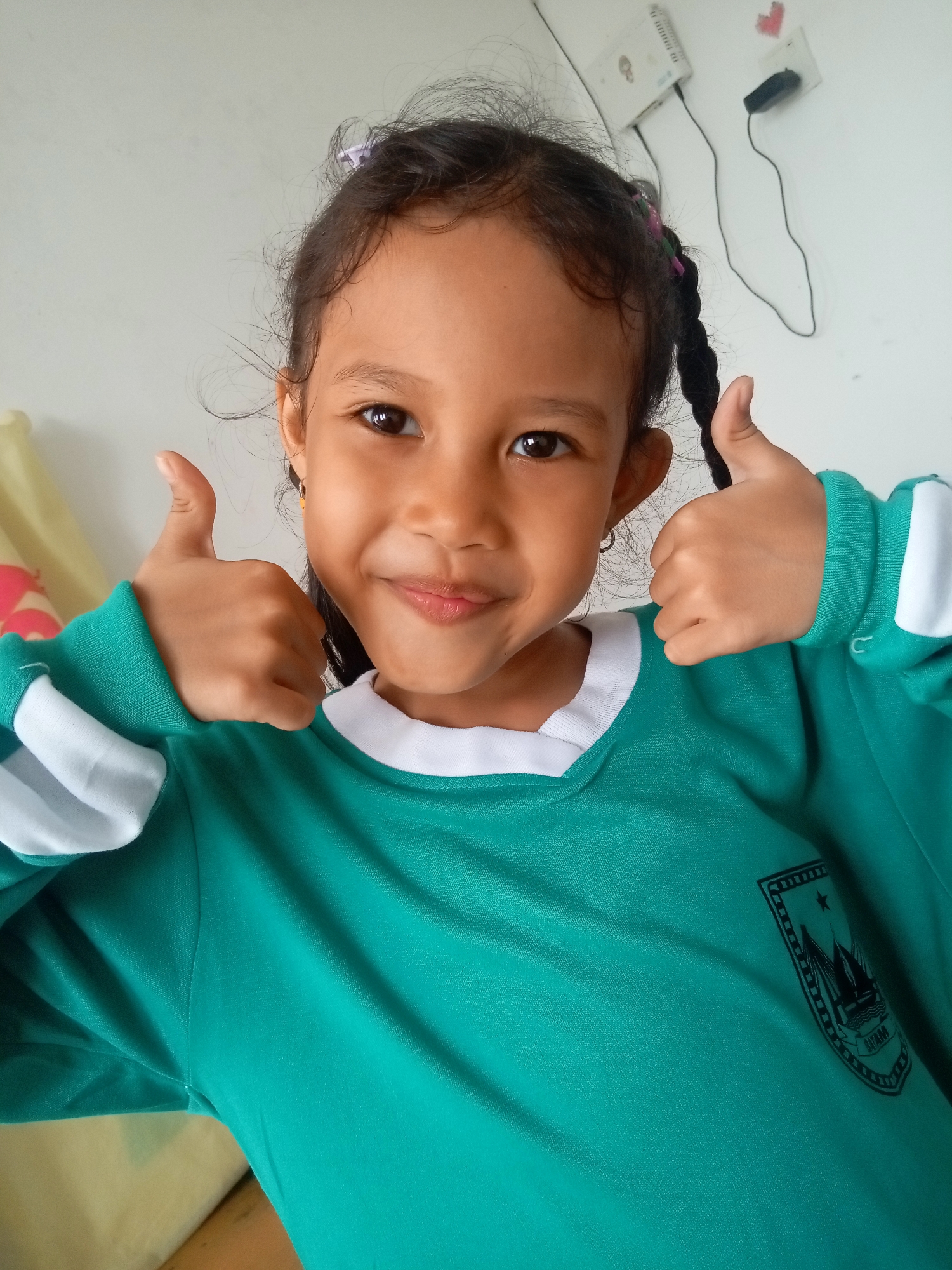 A young Indonesian girl with braided dark hair and a teal school uniform is pictured indoors, giving a double thumbs-up. She has dark eyes and a slight smile. The background is a light-colored wall with electrical outlets and cords. The overall impression is one of a happy and positive gesture. The image has no discernible features to indicate a specific location within Batam, Indonesia.