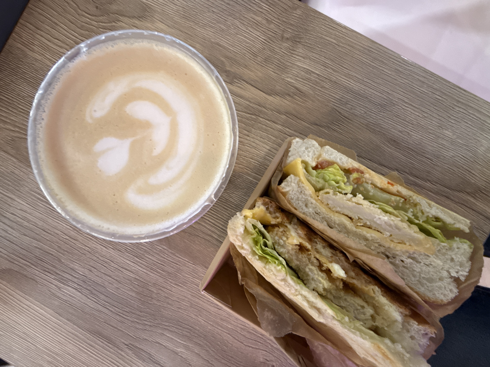 An overhead shot captures a takeaway meal set on a light-colored wooden table with horizontal grain. The scene is located in Moscow, Russia.
To the left, a clear plastic cup holds a coffee, likely a latte or cappuccino, distinguished by its light brown liquid topped with white foam. Intricate latte art, resembling a heart or a stylized leaf, adorns the surface of the foam, indicating careful preparation.
To the right, a sandwich, cut diagonally into two halves, is presented in a small, light brown cardboard tray or box lined with brown paper. The sandwich appears to be made with toasted or grilled white bread, possibly a type of baguette or ciabatta. Visible fillings include layers of cooked protein (likely chicken or a cutlet), melted yellow cheese, fresh green lettuce, and hints of red, possibly tomato or a sauce. The sandwich's packaging suggests it is prepared for takeout or a casual, on-the-go meal.
There are no people visible in the image, and no visible text on any of the items. The overall impression is a casual meal or snack, possibly a coffee break or lunch. The lighting suggests daytime.