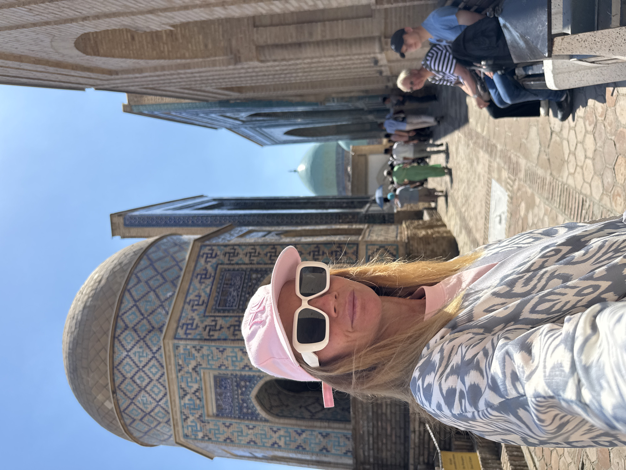The image shows a woman in sunglasses and a pink baseball cap, taking a selfie in front of a building with intricate blue tilework. In the background, there is a crowd of people walking along a stone-paved path with arched buildings on both sides. A large green dome is visible further in the background. The image was taken in Samarkand City, Uzbekistan.
