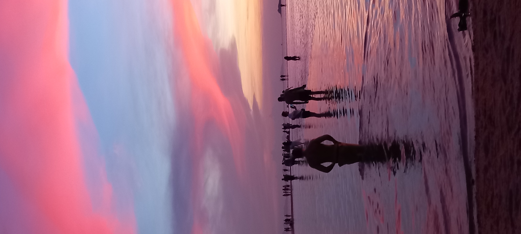 The image depicts a beach at sunset in Baan Sri Thanu, Thailand. Several silhouetted people stand in the shallow water, some appearing to be wading or swimming. The sky is a gradient of colors, ranging from pink and purple to orange. The wet sand reflects the colors of the sky. The beach is on the right. The people are silhouettes from the sunset.