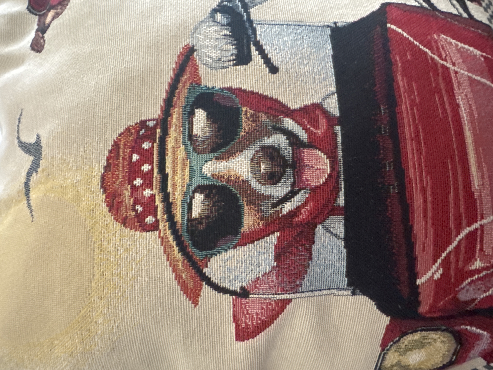 The image displays a close-up of a vibrant woven textile, likely a tapestry or cushion cover, featuring a stylized, anthropomorphic character. The character, oriented sideways in the frame, is depicted with a jovial expression, wearing a red, polka-dotted sombrero-like hat and what appears to be a red jacket or vest. Its face shows large, expressive eyes (one light blue/green, one brown) and a wide, dark smile, suggesting cheerfulness.
To the character's right (which is the top right in the rotated image), there is a large, red and black object resembling an accordion or another musical instrument, implying the character is playing music or performing. The texture of the textile is clearly visible, with distinct woven threads creating the image in various colors including reds, blues, greens, yellows, blacks, and creams, set against a light beige background. In the background, partially visible in the upper left, there's a hint of another figure or element, possibly a leg or boot. The overall aesthetic is festive and indicative of Mexican folk art, aligning with the cultural context of Puebla, Mexico. There is no visible text.