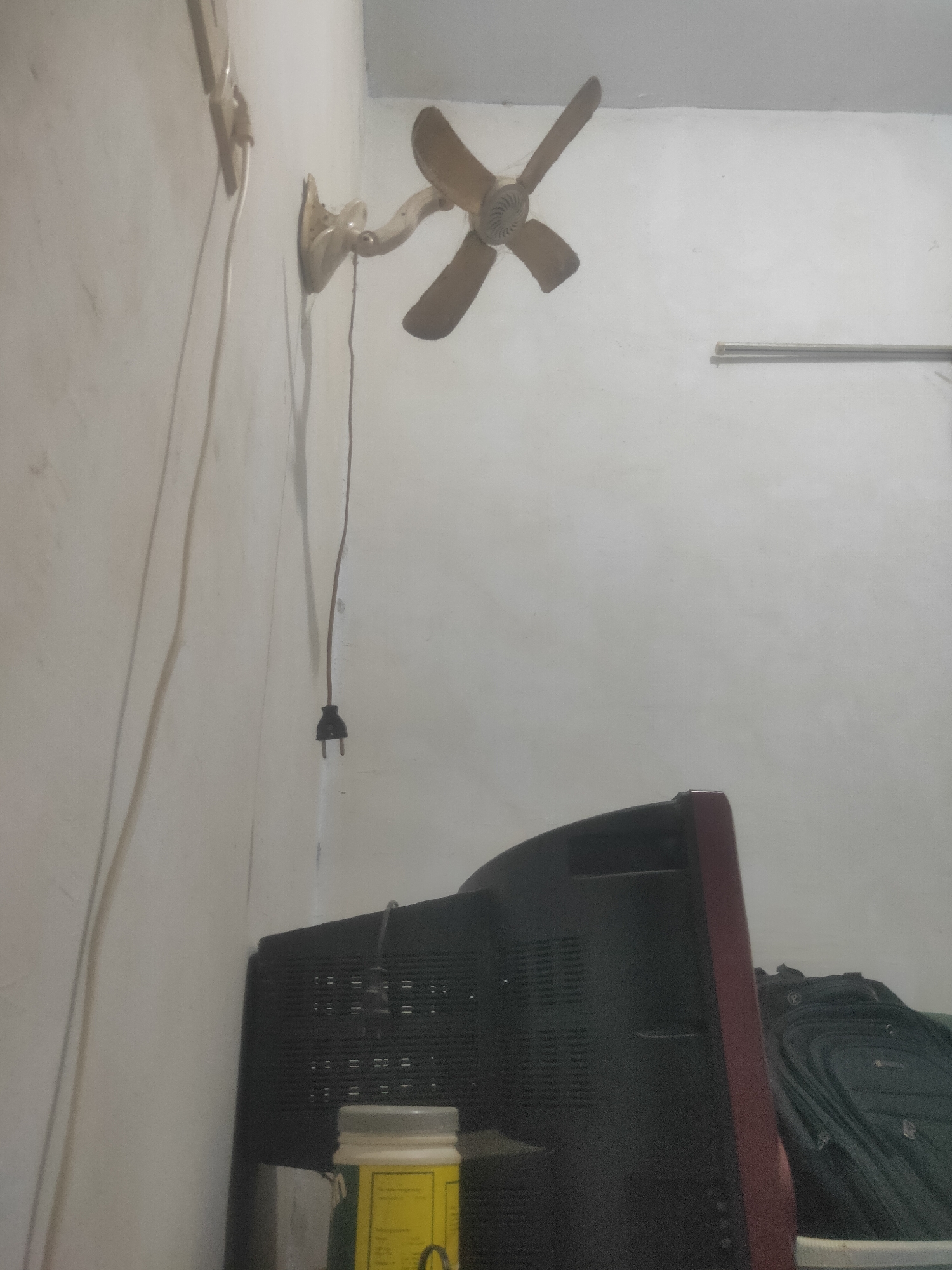 The image captures an interior scene within a room, likely a residential dwelling, in Antirogo, Indonesia. The primary subjects are a vintage wall-mounted oscillating fan and a television set. The fan, with its light-colored blades and housing, is positioned on a white wall, extending outwards with its articulated arm. A power cord for the fan is visible, leading down to an electrical plug dangling near the wall. To the right, a dark-colored television with a reddish trim sits on a surface, partly obscuring a dark green backpack. The wall surface appears textured and slightly aged, with a thin horizontal pipe or conduit running across it in the background. The lighting suggests an indoor environment, with soft illumination casting subtle shadows. No individuals are present in the frame, and the scene is static, depicting everyday objects in a typical domestic setting in Antirogo.