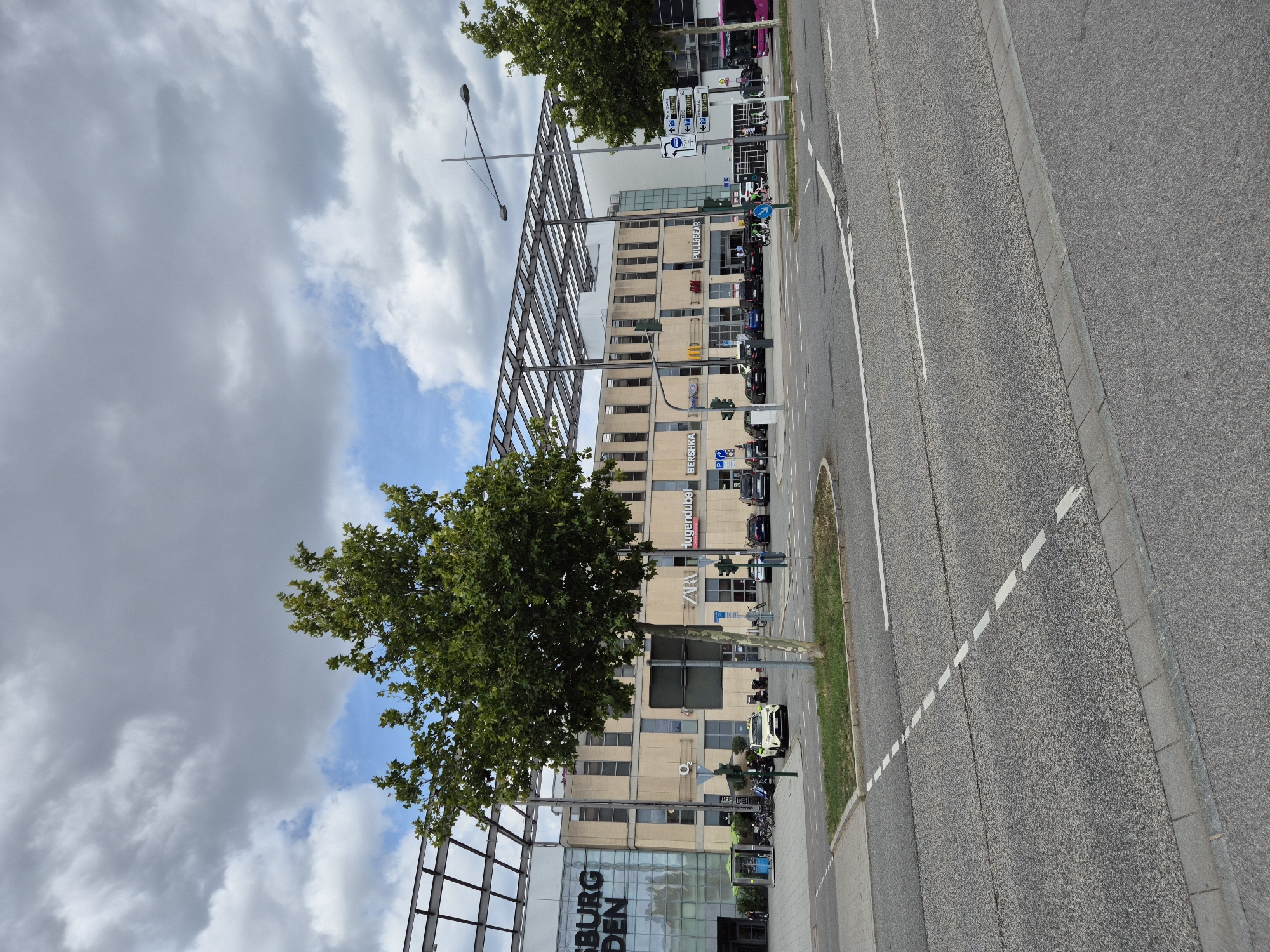 A wide shot captures a street scene in Regensburg, Germany, with a modern shopping complex dominating the background. The building features a glass facade with the words "SÜDBURG" and "DEN" visible. Several retail storefronts are present, including "ZARA", "HUGENDUBEL", and "PULL&BEAR". A white police car is parked in front of the complex, and several pedestrians are visible walking near the entrance and riding bicycles and e-scooters. The street is paved and has clear lane markings, and a traffic light is visible. Trees line the sidewalk, and a large cloudy sky fills the upper portion of the image. The scene appears to be a typical day in a European urban environment.