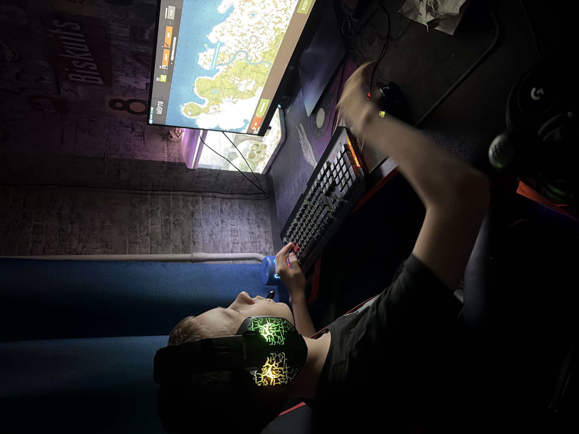 The image displays a dimly lit room in Chaikovsky, Russia, occupied by a young male engaged in computer gaming. A fair-skinned male, appearing to be a pre-teen or teenager, is seated in a dark gaming chair, oriented towards a computer monitor positioned to his right. He wears a black short-sleeved t-shirt and black over-ear gaming headphones featuring illuminated green and yellow lightning-like patterns, with the "SVEN" brand visible.
His right hand operates a black gaming mouse with a glowing red palm print emblem, resting on a large dark mousepad. His left hand is positioned over a black mechanical keyboard, which exhibits red and orange backlighting along its base and keys. The computer monitor displays a digital topographical map with landmasses, water bodies, and various markers. Text visible on the monitor includes "МЁРТВ" (DEAD) in the top left corner, and a numerical value of "22.8м" in the top right.
Adjacent to the left side of the monitor is a small, rectangular fish tank containing a white skull decoration and emitting a purple internal light. The room's background features a dark wall with a distinct brick-like texture. Mounted on this wall, above the monitor, are various signs, including "Biskuiits" in red lettering and "Blade" (partially obscured), alongside large white numbers "8" and "4". To the left of the frame, dark blue vertical curtains with a textured weave are visible. A small, blue cylindrical device is located on the desk between the male and the curtains. A separate black Logitech brand headset is present on the desk surface in the lower right portion of the scene.