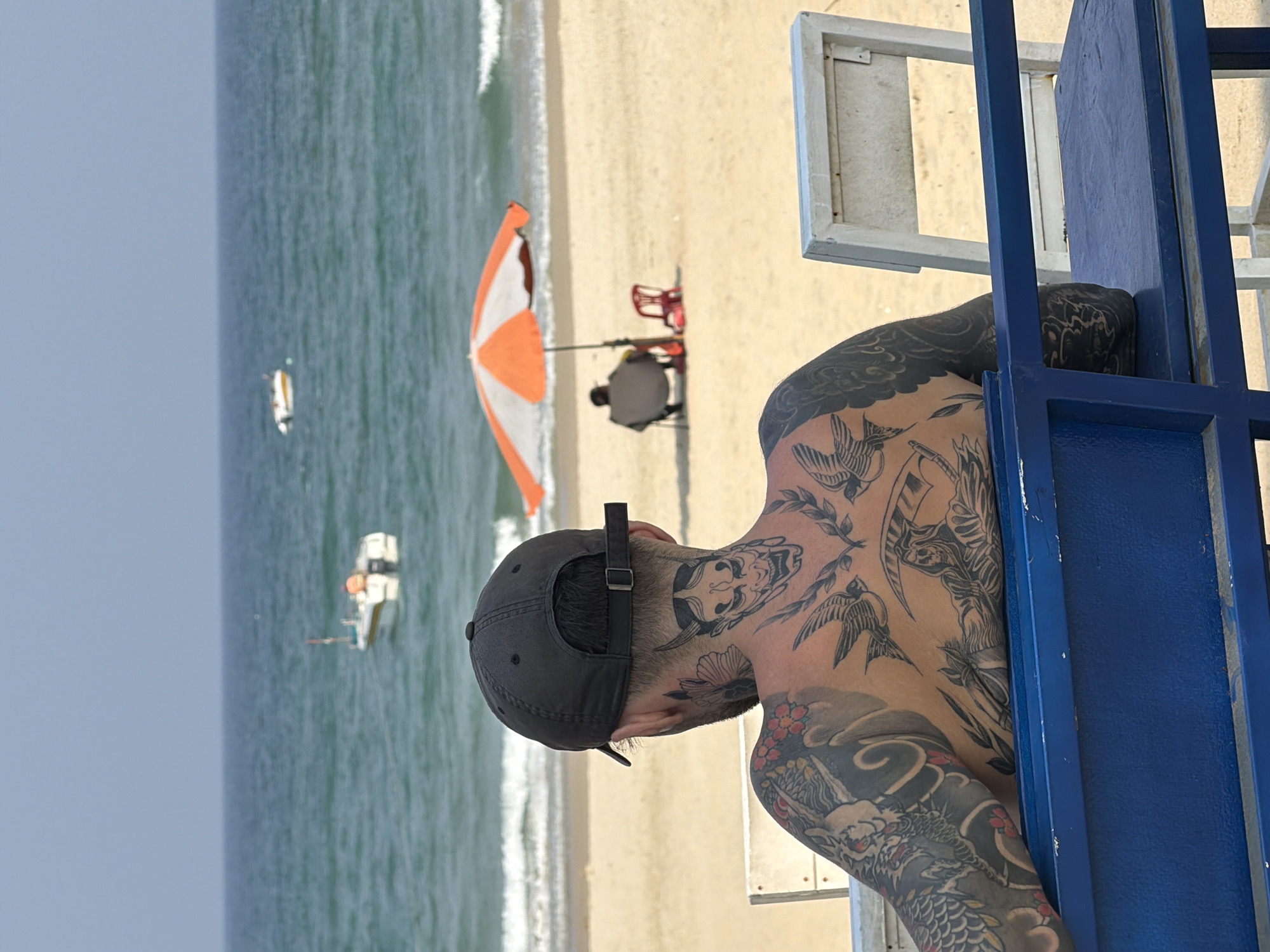 The image depicts a man with extensive tattoos standing near a blue structure, possibly a lifeguard stand, at a beach in Đà Nẵng, Vietnam. The man is wearing a black baseball cap. In the background, the beach slopes down to the ocean, where two small boats are visible. Further along the beach, there is a person sitting under an orange and white umbrella next to a red beach chair. The scene appears to be taken during daylight hours.