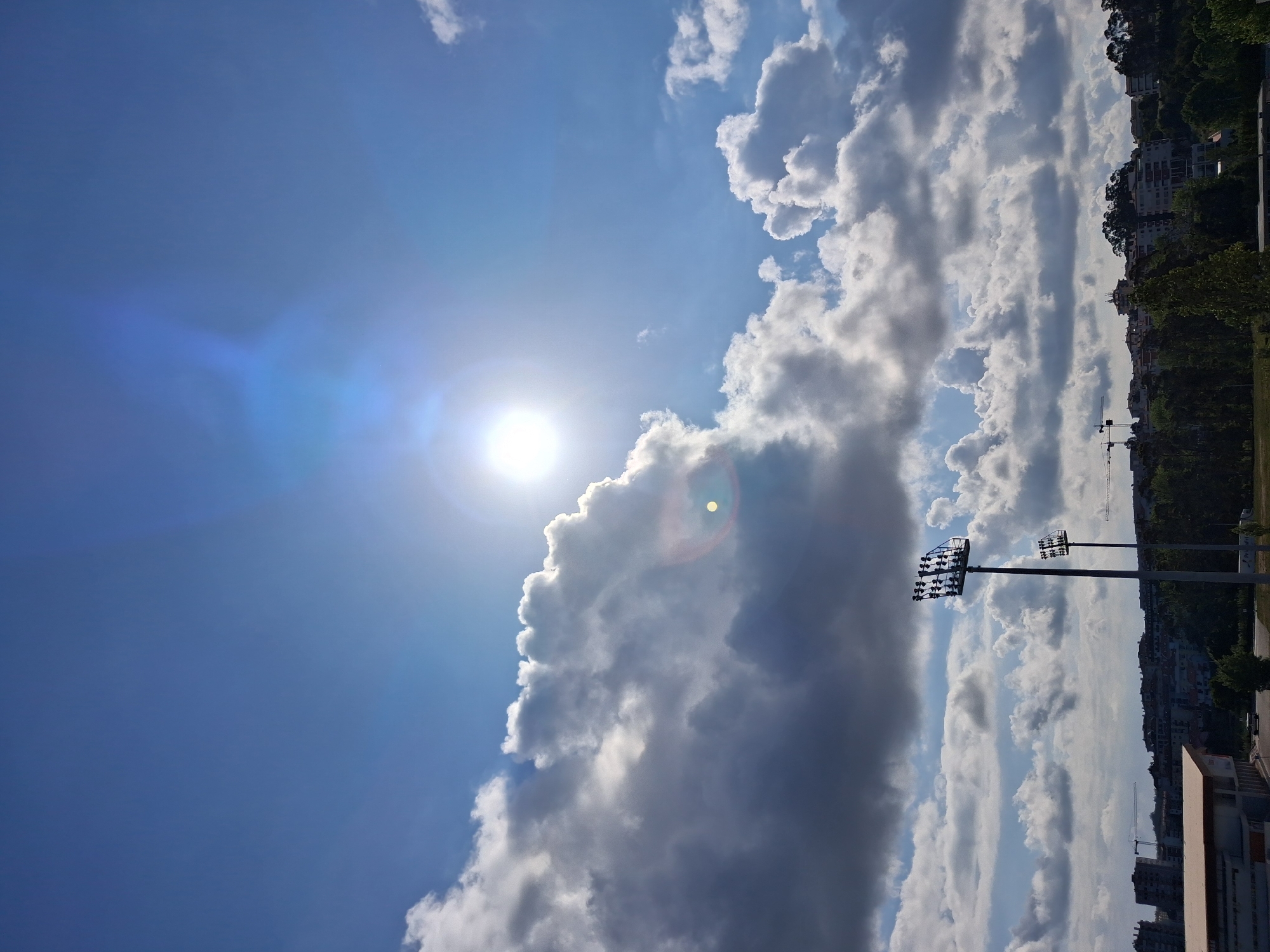 The image shows a bright sunny sky over Figueira da Foz, Portugal. Dominating the scene are cumulus clouds, both illuminated by the sun and casting shadows. The sun itself is high in the sky, causing lens flare. In the background, a portion of the cityscape is visible, including trees, buildings, and street lights. The overall scene is outdoors during daylight hours. The sky is blue.