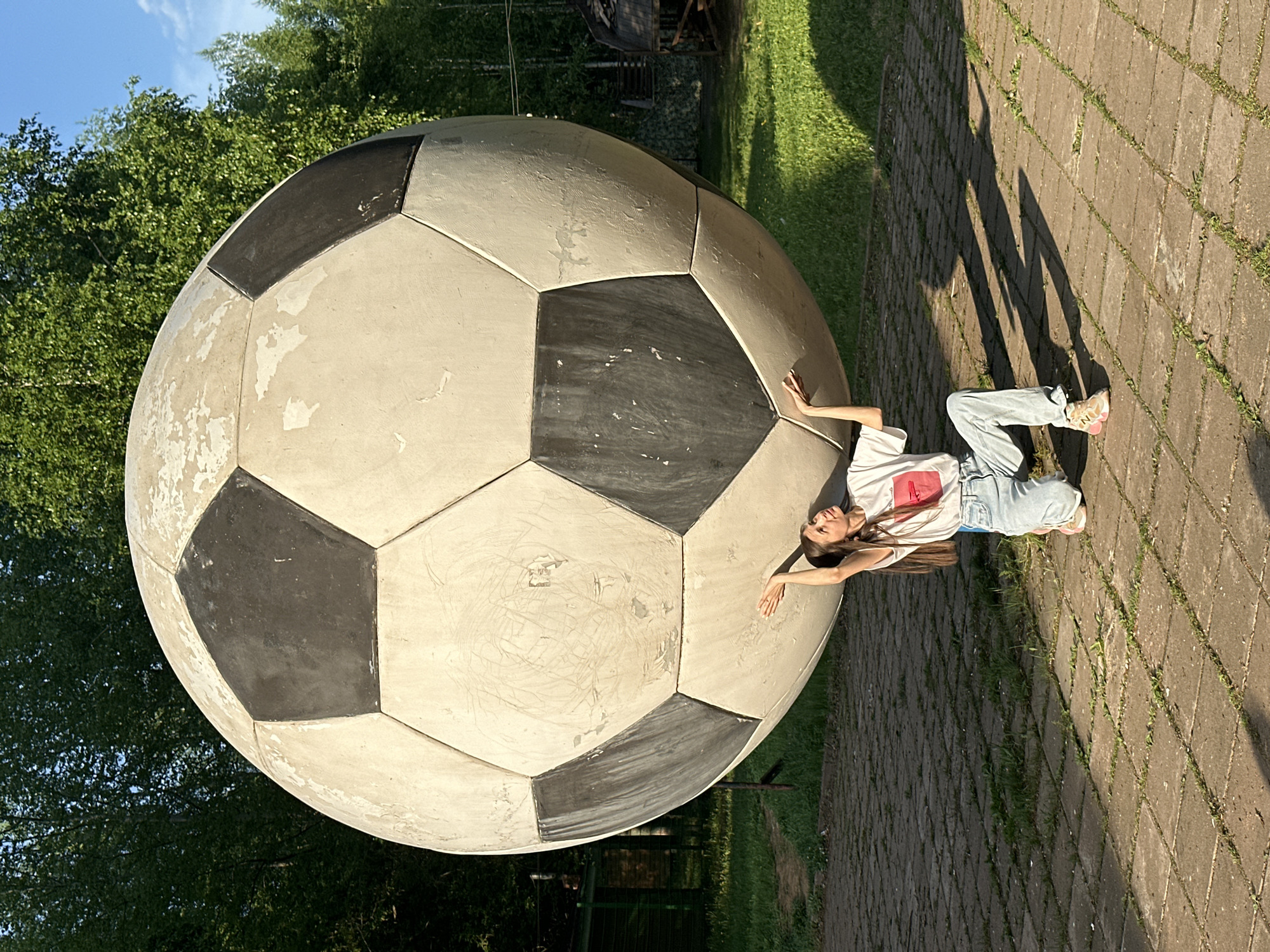 A low-angle, outdoor shot shows a large soccer ball statue with a girl posing beside it in Izhevsk, Russia.
The soccer ball is composed of white and black polygonal panels and has visible wear and tear. The girl is positioned lying down on the right side of the soccer ball, her head closest to the ball, arms outstretched. She is wearing a white T-shirt with a red graphic, light blue jeans, and sneakers.
The setting appears to be a park or recreational area, with paving stones forming a walkway on the right. Patches of green grass are visible in the background behind the soccer ball, along with trees, some of which are out of focus. A shadow cast by the girl and the large soccer ball is visible on the paving stones. The sky is partly visible in the upper left corner.