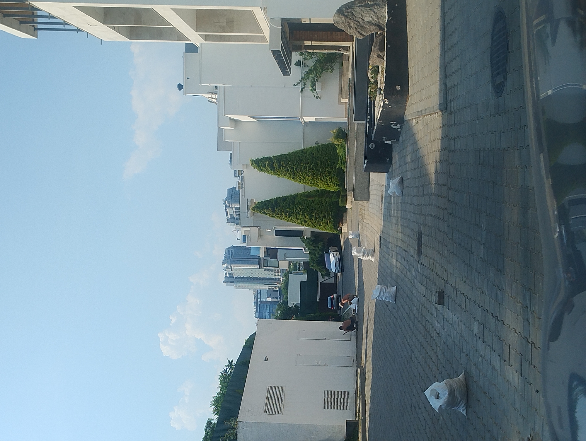 An outdoor, medium shot captures a paved area with several white sacks of material scattered on the ground. In the middle ground, two individuals are crouched down, appearing to be working with one of the sacks. Behind them, a dark-colored sedan is parked, and a white car is driving away. The background reveals modern, multi-story white buildings and some taller city buildings in the distance. Two conical evergreen trees flank the white buildings on the right. The sky is clear blue with a few scattered white clouds. The scene is illuminated by daylight, casting shadows on the paved surface.