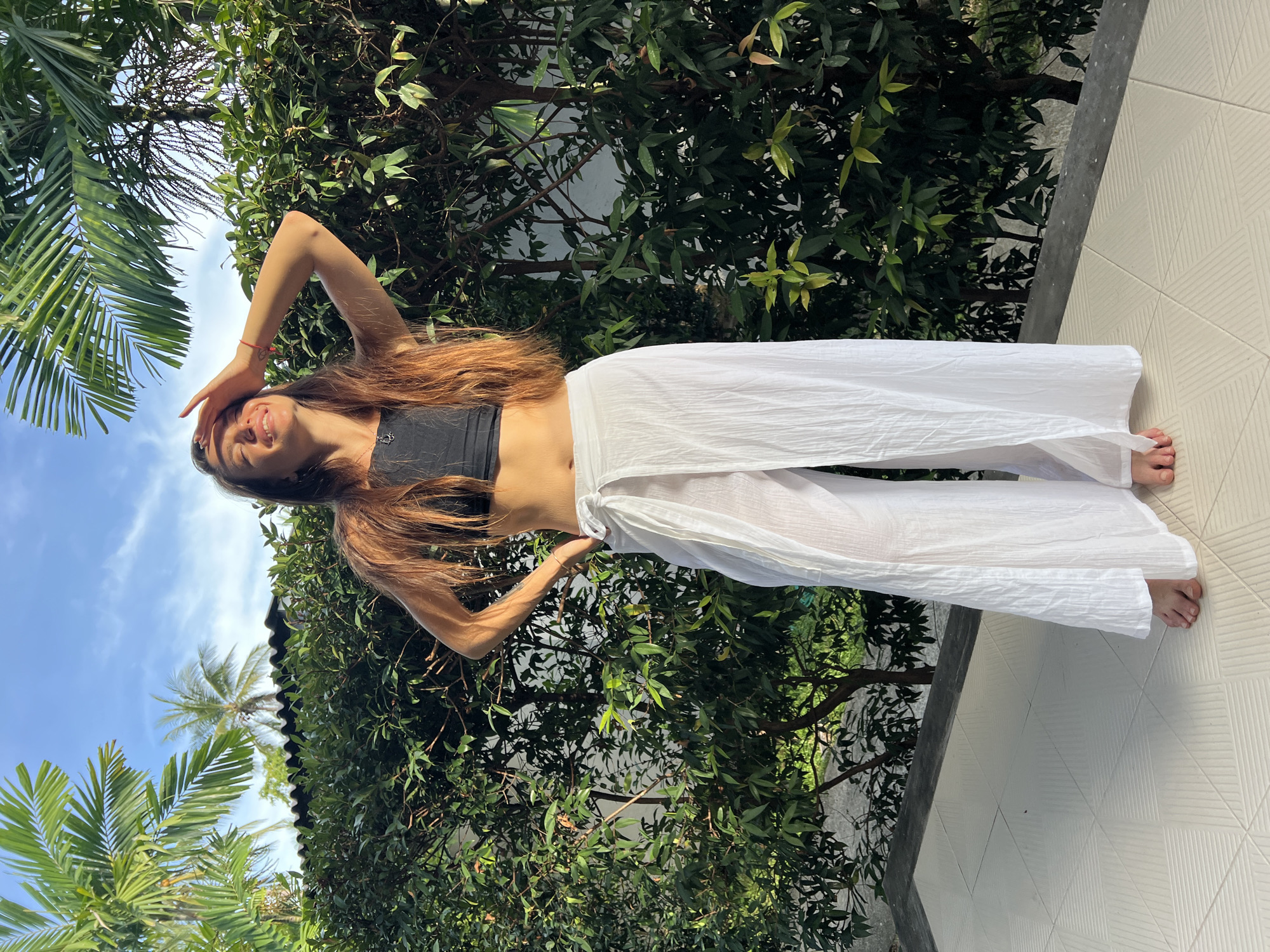 A young woman with long brown hair is posing outdoors in บ้านบางรักษ์, Thailand. She wears a black crop top and white pants, with one hand on her head and the other on her hip. Lush green foliage and palm trees form the background, with a glimpse of blue sky. The woman stands on a tiled surface.
