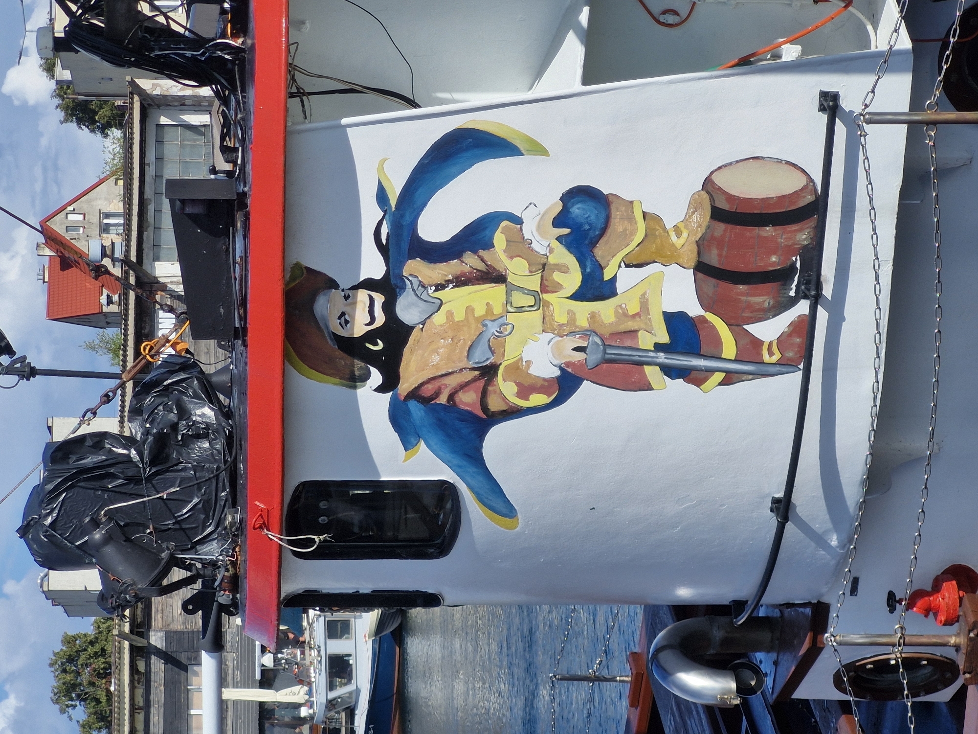 The image shows the side of a white boat with a red trim, decorated with a painted pirate figure. The pirate is depicted in a brown hat, yellow and brown clothing, and a blue cape, standing on a red barrel. The boat is docked in a harbor, with other boats and buildings visible in the background. The water is blue and calm. The scene is likely located in the city of Hel, Poland, known for its maritime history and numerous boats. The painting on the boat is a whimsical representation of a pirate, adding a touch of character to the vessel.