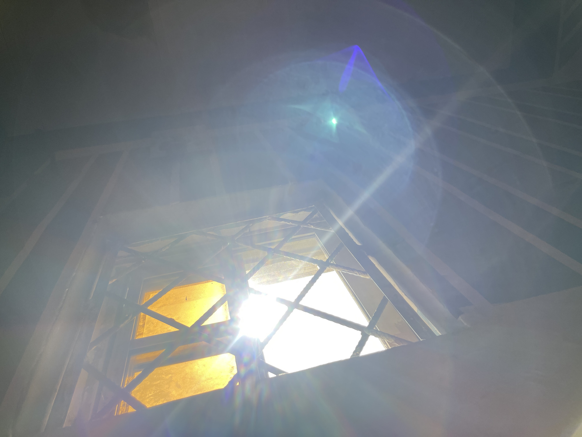 The image depicts a view looking upwards through a barred window, with bright sunlight streaming through. The bars form a diamond pattern across the window. The light is so intense that it causes lens flare and glare, with a rainbow-like effect visible on the left side and a bright blue and green flare on the right. Behind the bars, there appears to be a yellowish translucent material, possibly a curtain or shade. The surroundings are dimly lit and out of focus, suggesting an indoor environment. The scene suggests an exterior view through a protected opening, possibly from a ground-level or lower-floor building looking up towards the sky. The image does not provide specific details that would allow for precise localization within the city of Rafin Par, Nigeria, but it captures a common architectural feature and lighting condition.