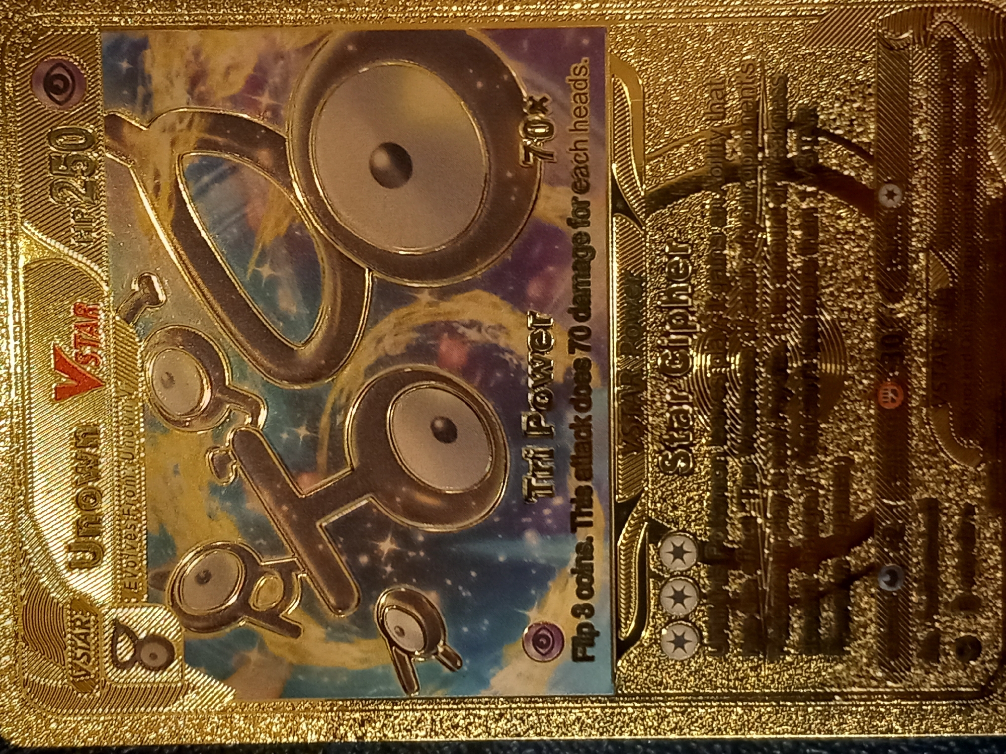 This is a close-up, angled shot of a gold Pokémon trading card. The card features a Pokémon with a metallic, round head and body, depicted against a starry, galaxy-like background. The Pokémon has a unique symbol for a face. On the left side of the card, text reads "VSTAR UNOWN VSTAR 250" and "Evolves From Unown V". The center of the card displays the Pokémon's attack name, "Tri Power," with the damage output of "70x" and a description: "Flip 3 coins. This attack does 70 damage for each heads." To the right, more text is visible, including "STAR CIPHER," and "ABILITY THA". The entire card is encased in a textured gold border, with gold embellishments and patterns. The lighting suggests it's an indoor setting, likely highlighting the collectible nature of the card. There are no people or animals visible.