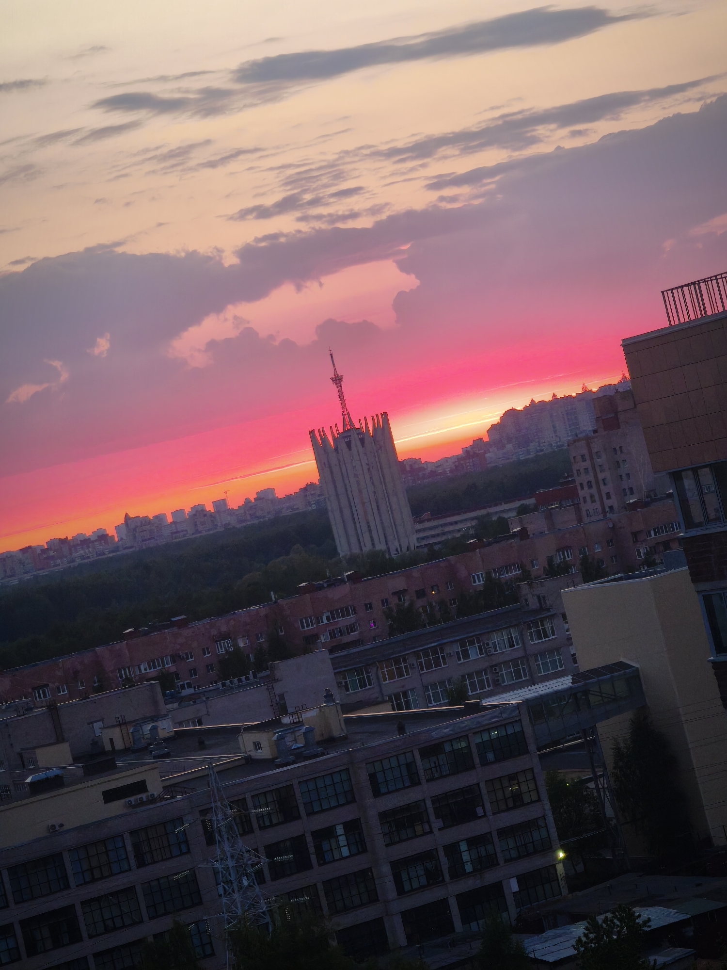 The image depicts an urban landscape in Saint Petersburg, Russia, during a vibrant sunset. In the foreground, a cluster of multi-story buildings with visible windows, some connected by enclosed walkways, suggest an industrial or residential area. Beyond these buildings, a dense green belt of trees stretches towards the horizon. Further back, a row of taller buildings forms the cityscape, silhouetted against the bright, colorful sky. The sky transitions from a bright pink-red near the horizon to lighter hues of orange, pink, and grey at the top, with scattered clouds adding texture. A prominent architectural landmark with a central tower stands out against the sunset, suggesting a significant building within the city. The image is taken at an oblique angle, giving a sense of depth and perspective.