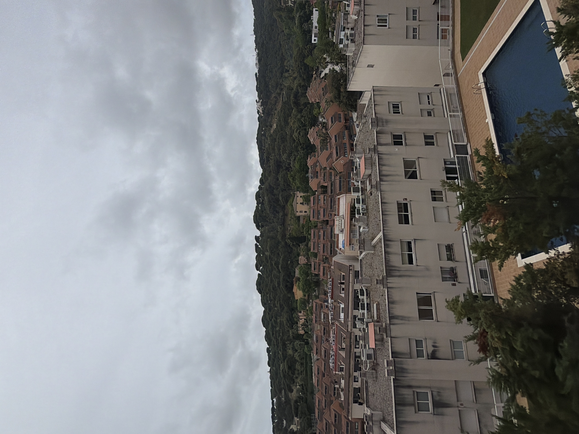 The image captures an elevated view in Sant Andreu de Llavaneres, Spain, under a cloudy sky. The scene encompasses dense forestation that ascends a hill, adjacent to residential buildings. A multi-story structure with numerous windows is prominently featured. A blue swimming pool is visible.